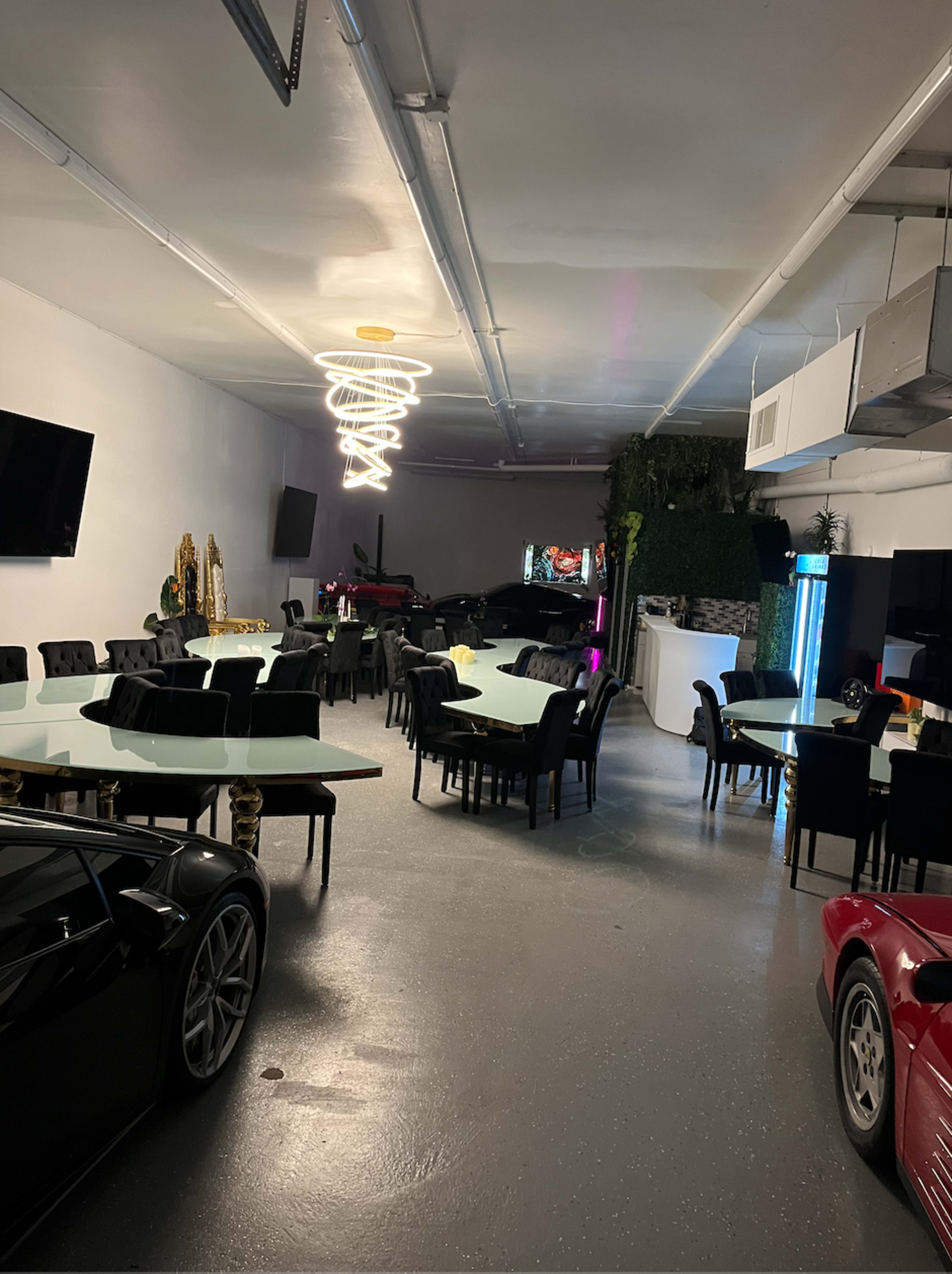 The image shows a spacious interior resembling a lounge area with multiple black dining chairs and tables, along with luxury cars displayed nearby.
