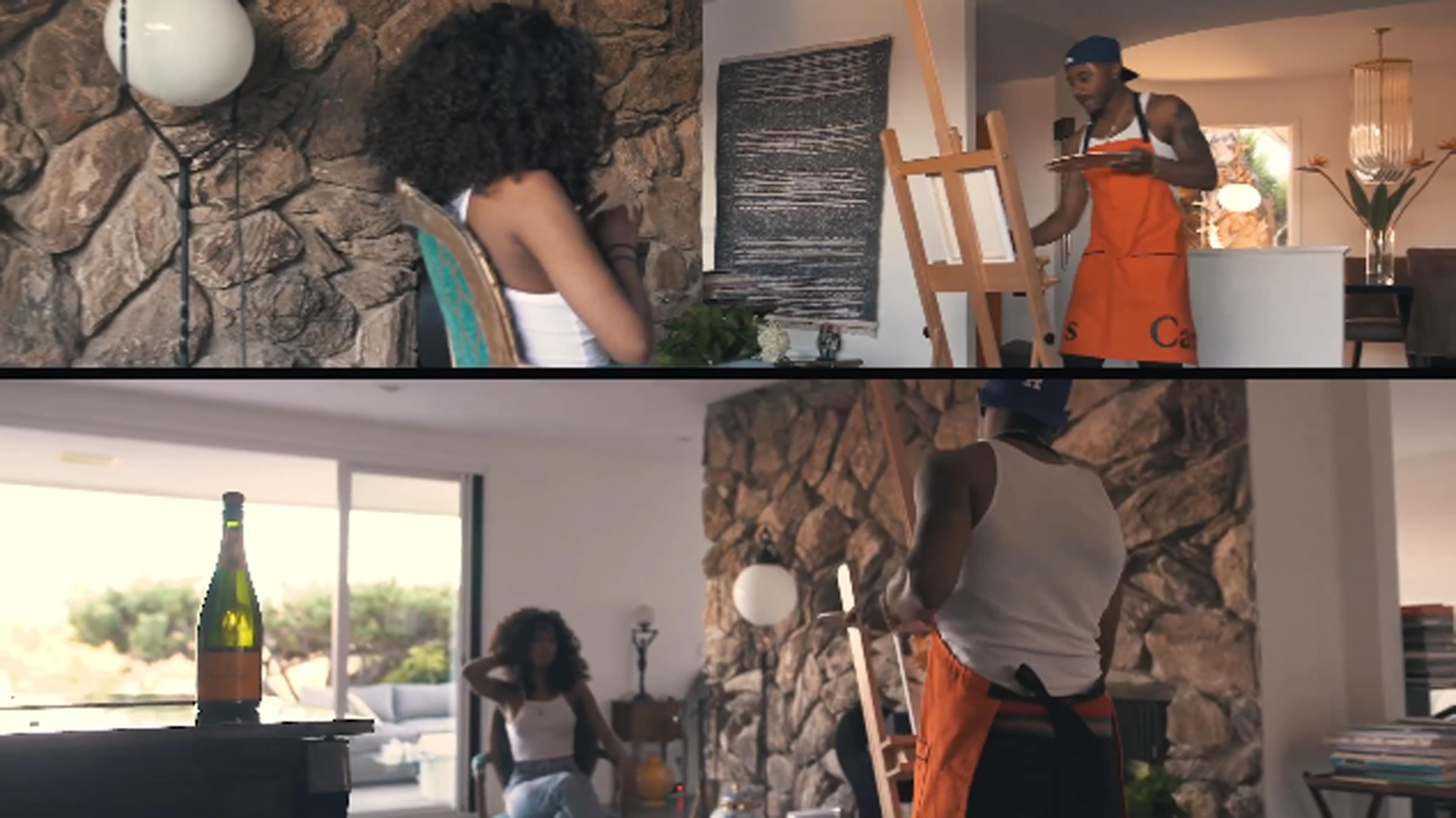 A woman sits in a chair while a man holds an easel, both in a room with a stone wall and large windows.