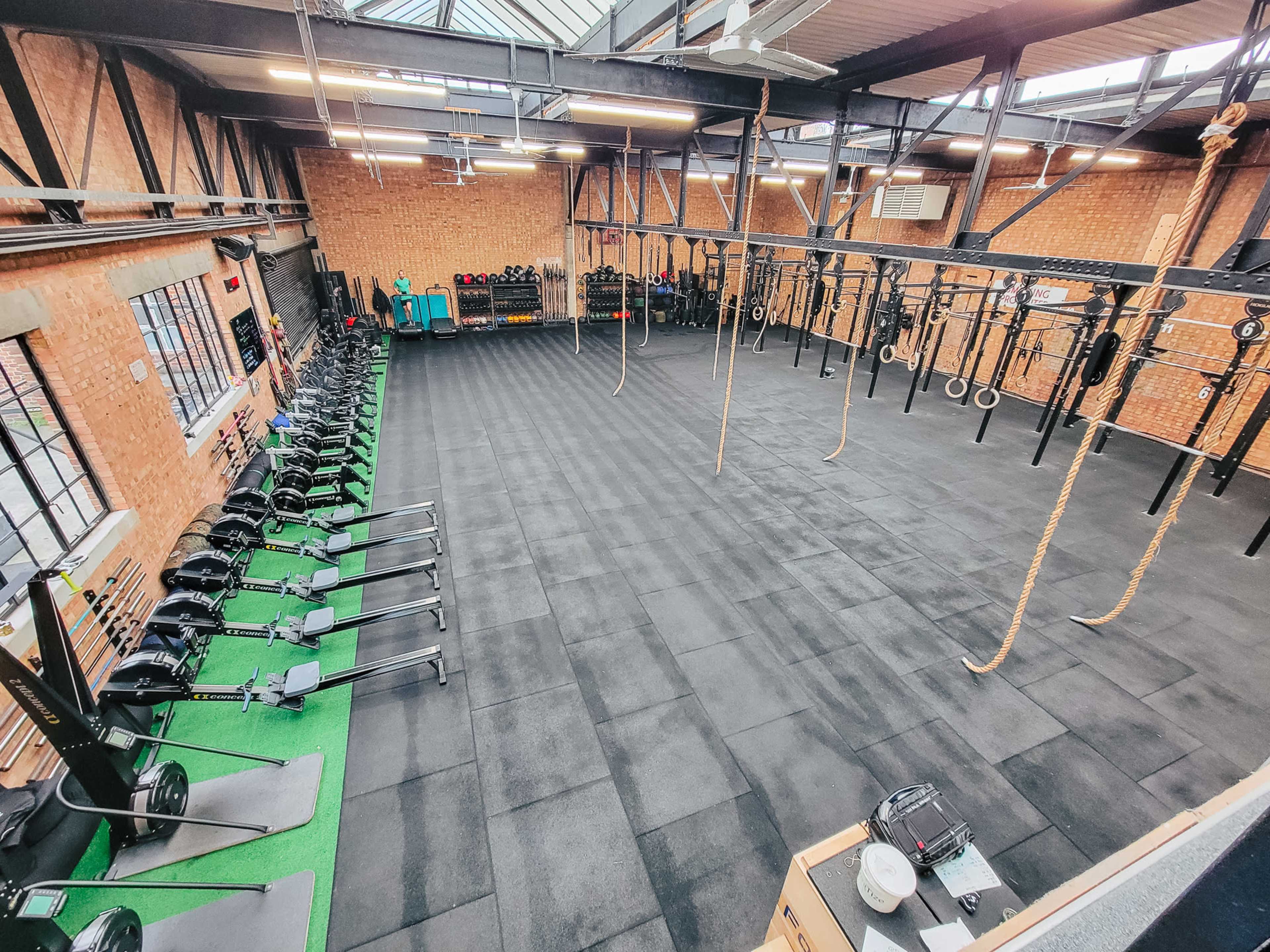 The image shows a spacious gym with exercise equipment, including rowing machines, suspended rings, and ropes, arranged on a rubberized floor with exposed brick walls.