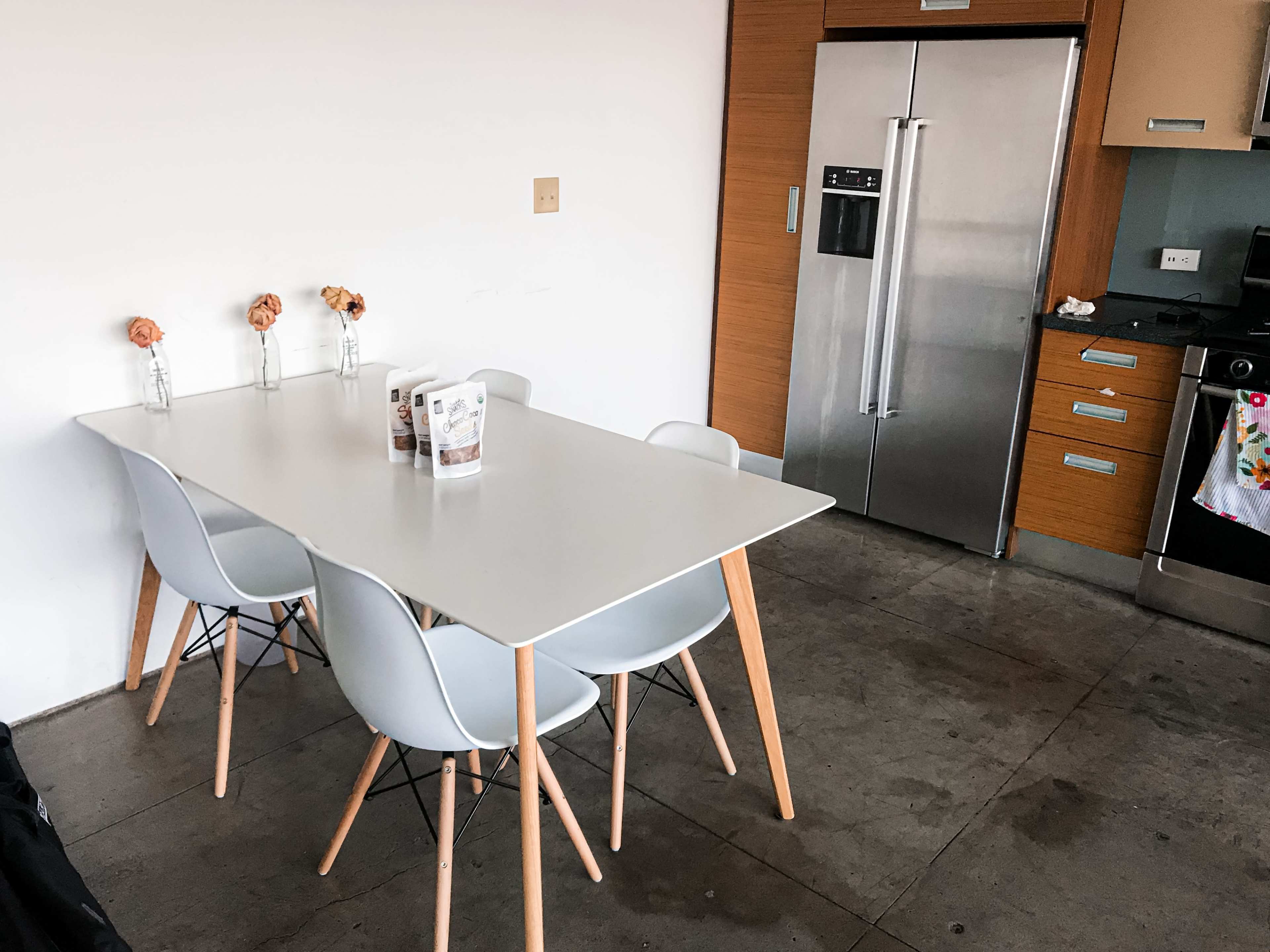A modern kitchen features a white table surrounded by four gray chairs, with a stainless steel refrigerator and a stove in the background.