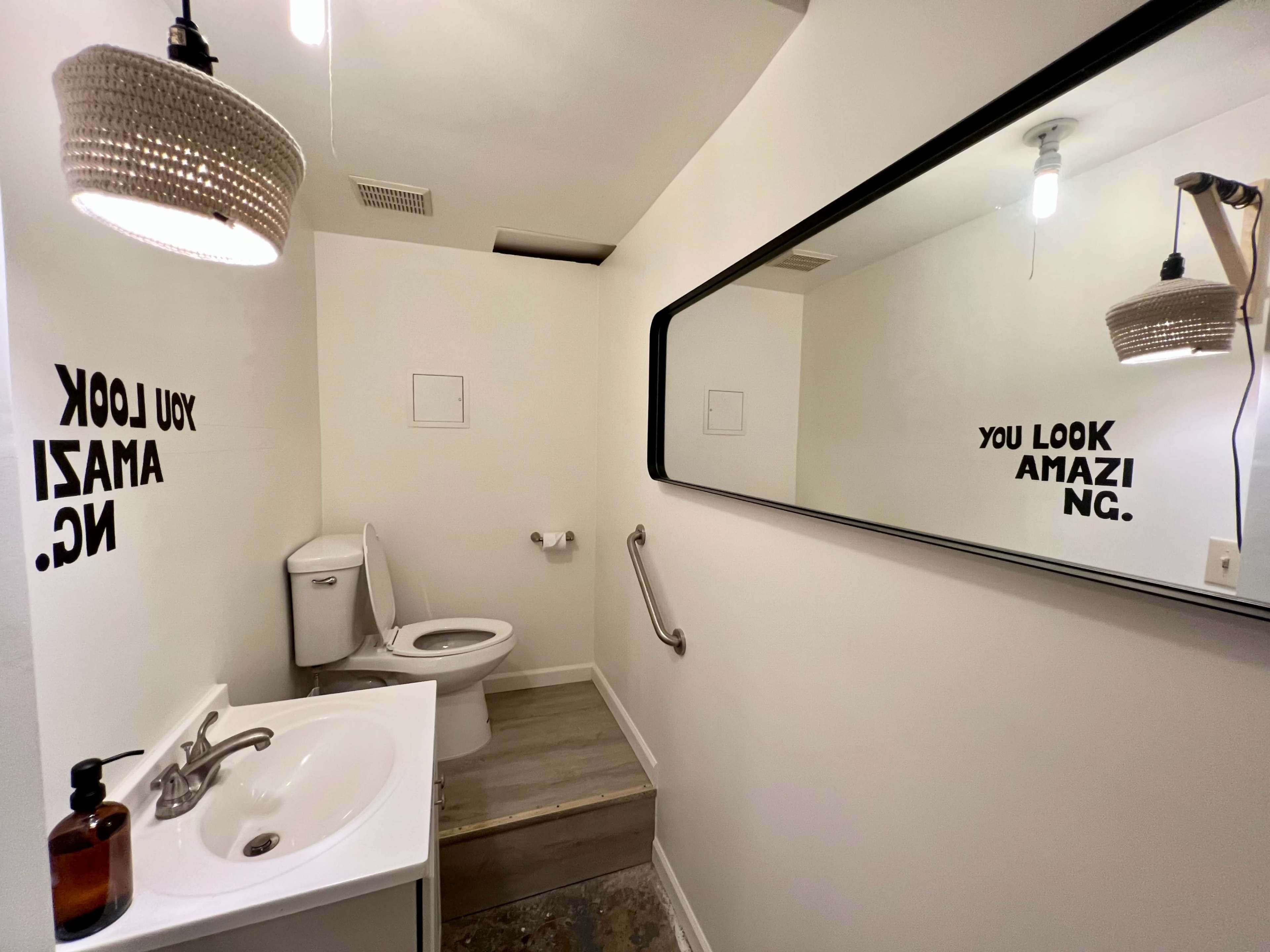 A small bathroom featuring a toilet, a sink with a soap dispenser, and a mirror with motivational text on the walls.