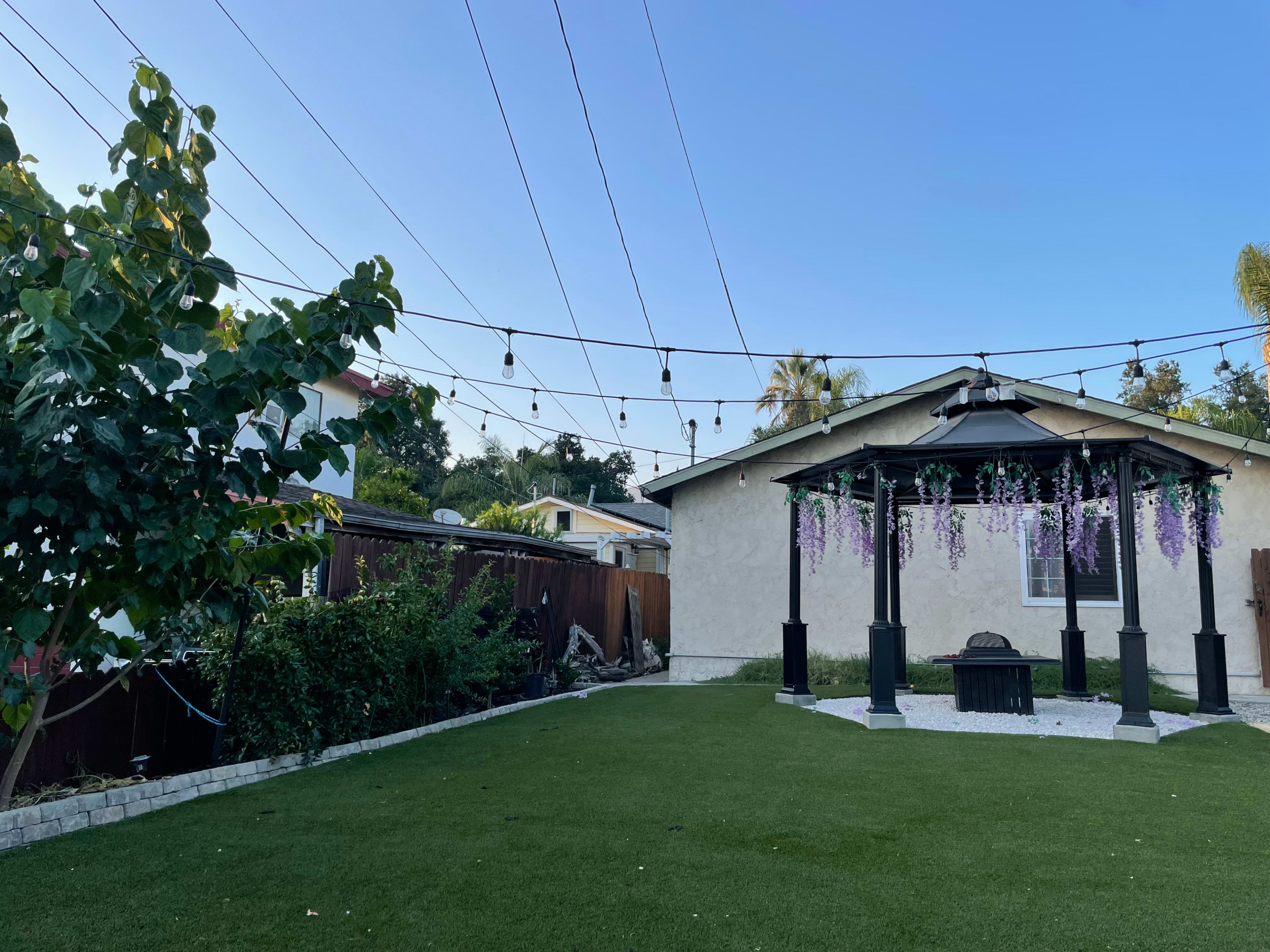 A backyard with artificial grass, a gazebo adorned with hanging flowers, and overhead string lights against a clear blue sky.