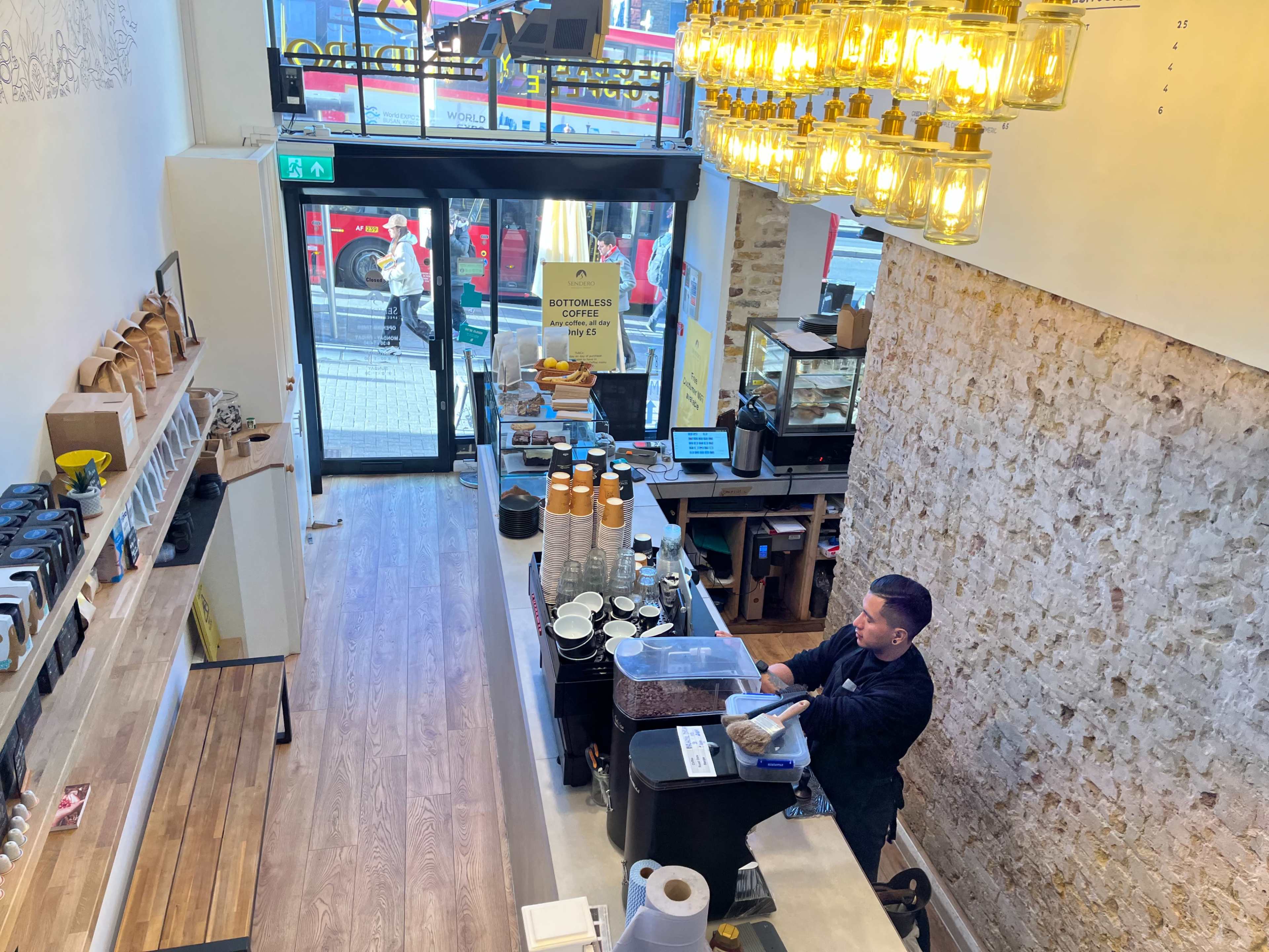 A barista prepares coffee at a café with large windows and decorative lighting, showcasing a busy street outside.