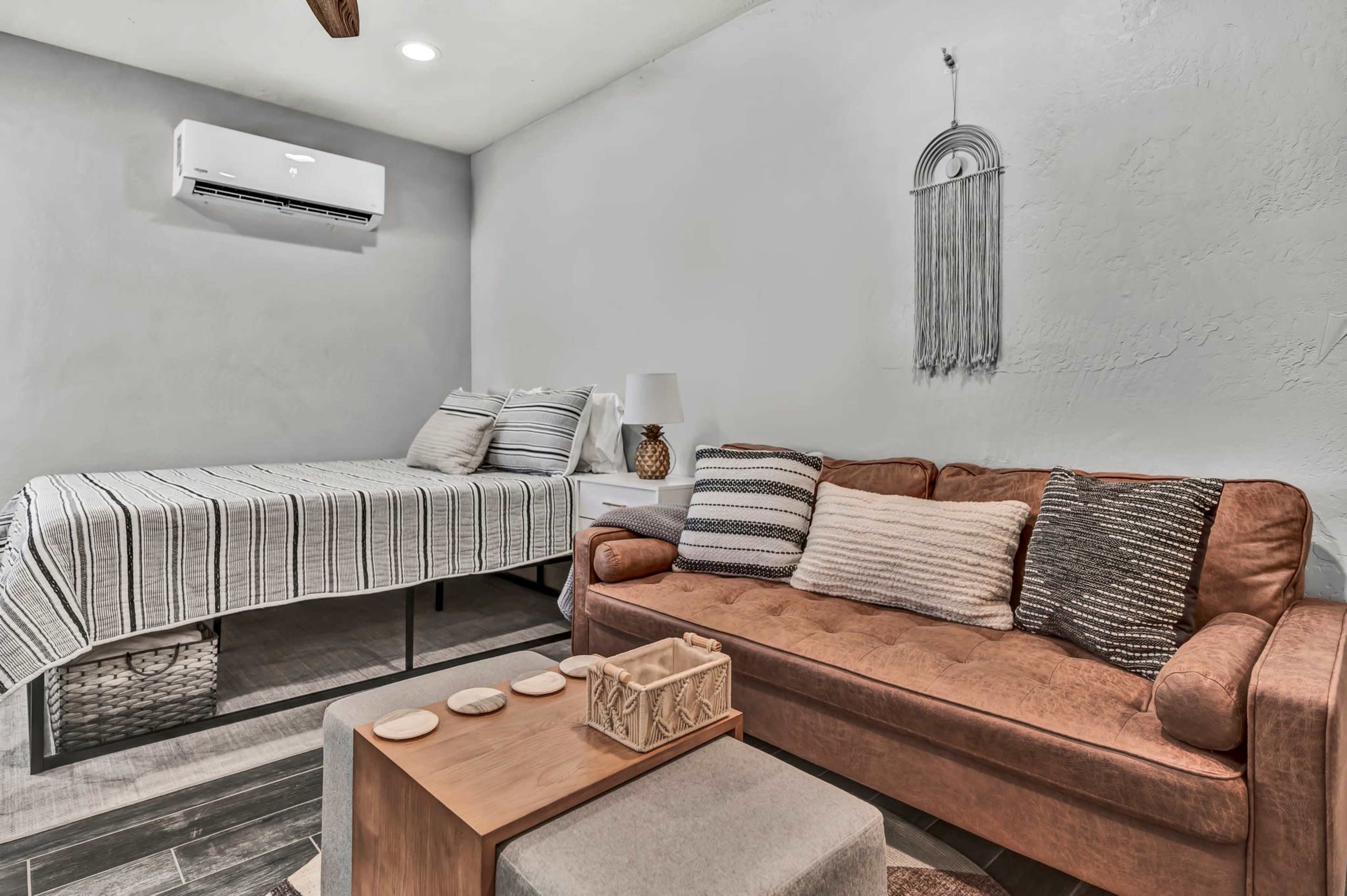 A small, cozy room featuring a bed with striped bedding, a brown sofa with decorative pillows, and a coffee table with a woven basket beside it.