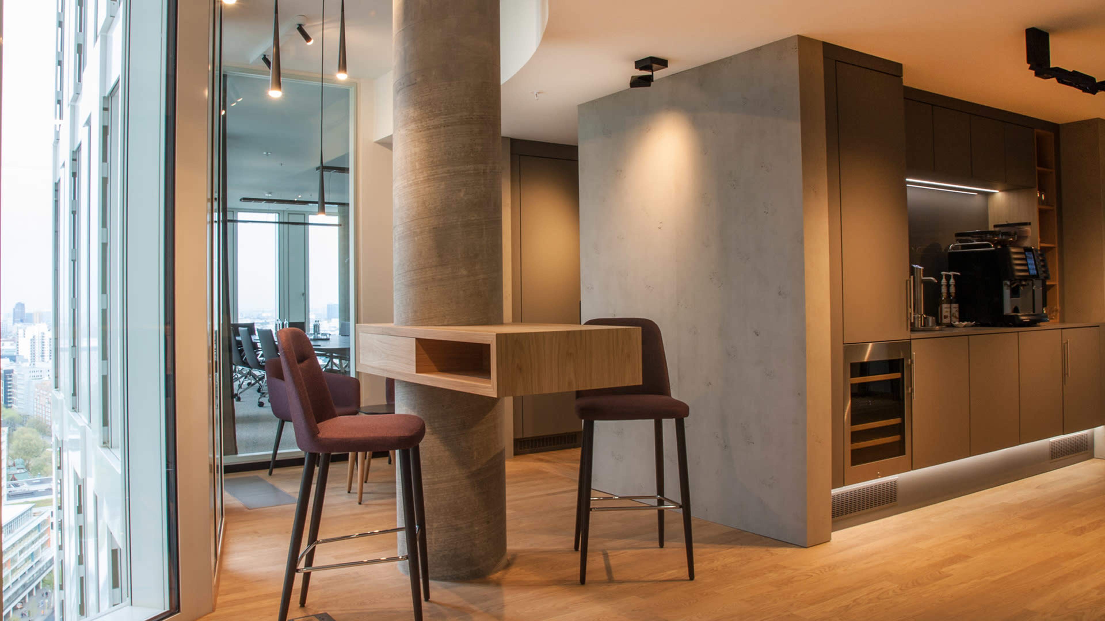 A modern office space featuring a small kitchen area with two high stools and a large window overlooking a cityscape.