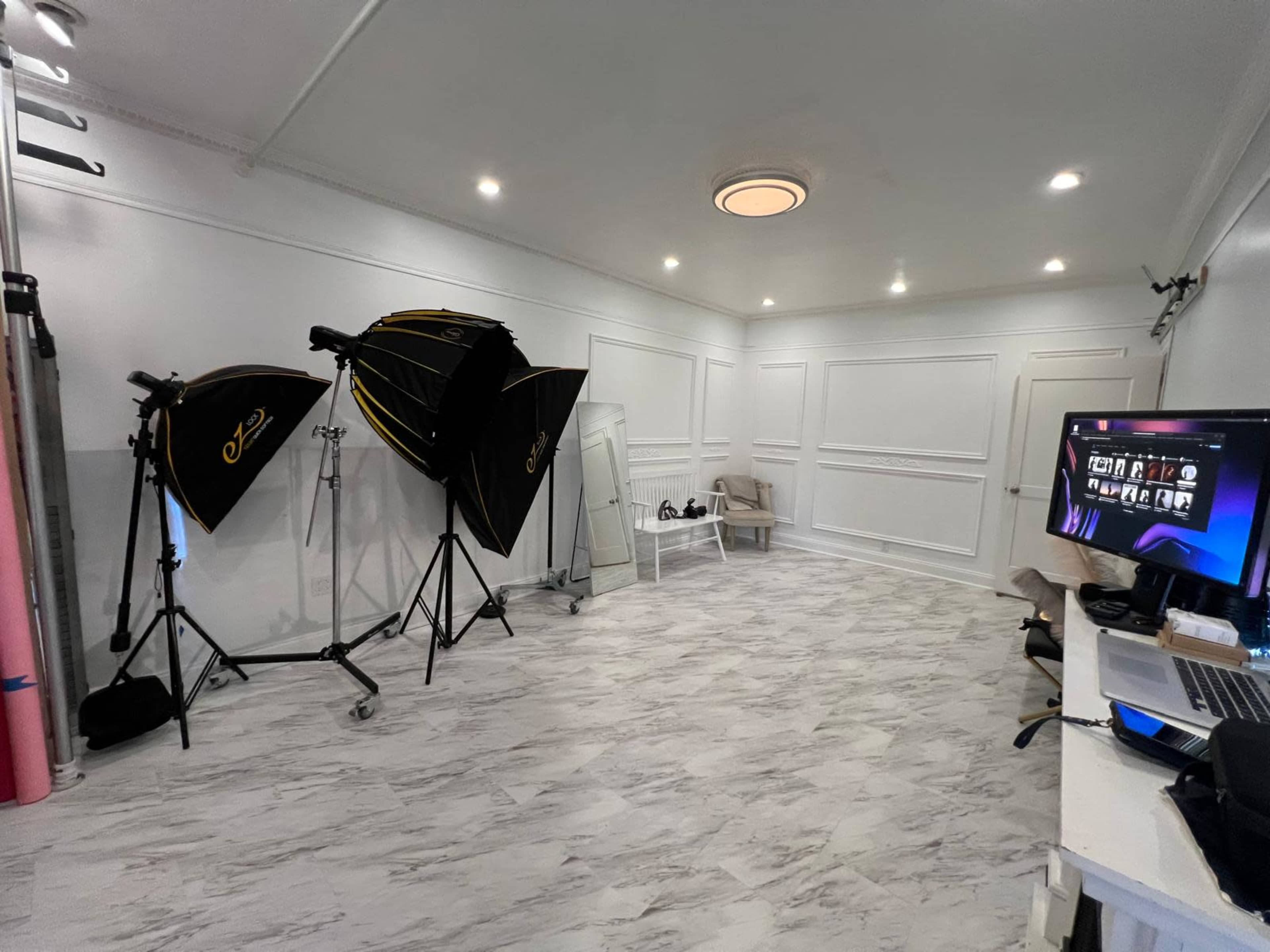 The image shows a well-lit photography studio featuring multiple softboxes, a mirror, and a desk with a laptop.