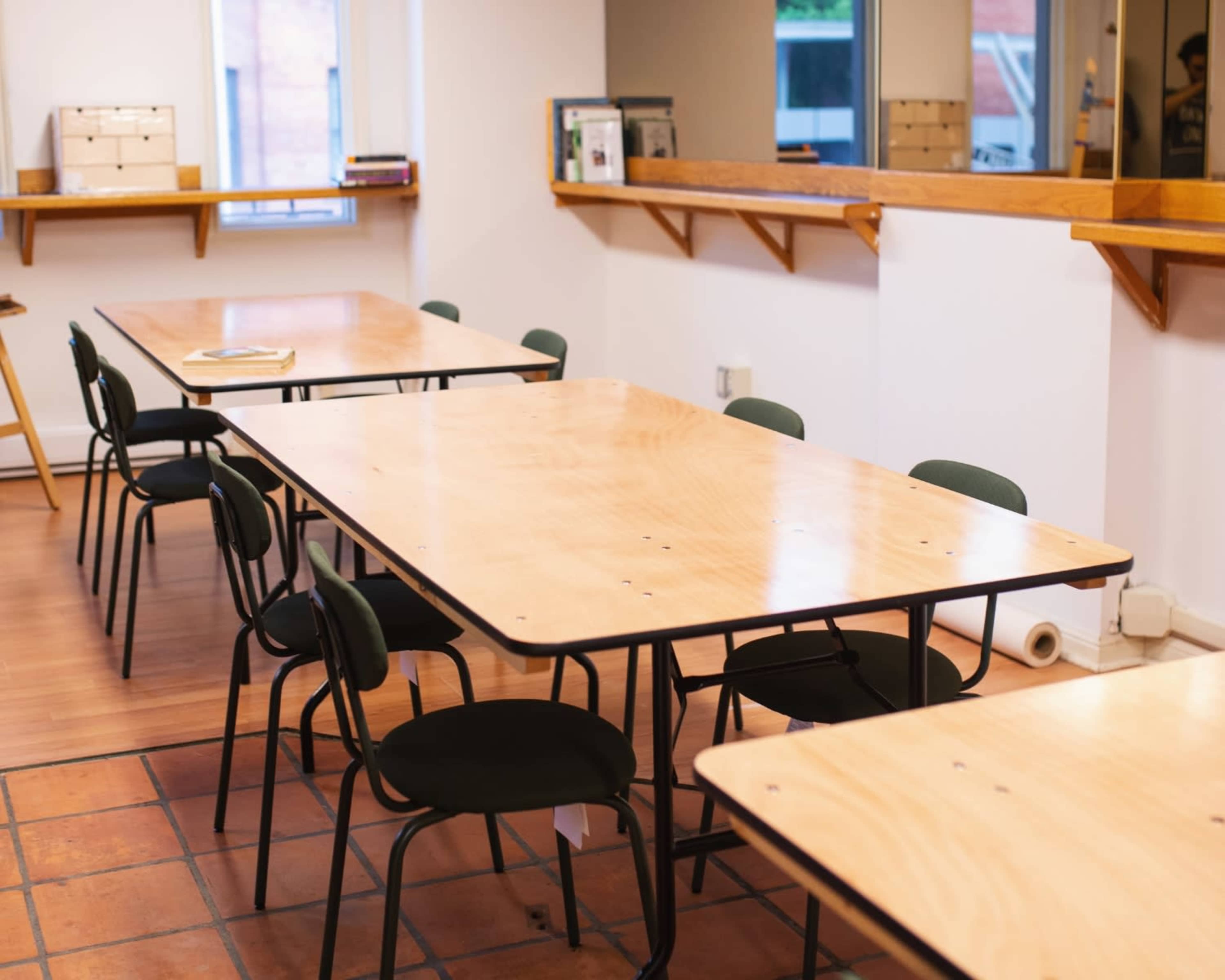 The image shows a classroom with several wooden tables and black chairs arranged around them.