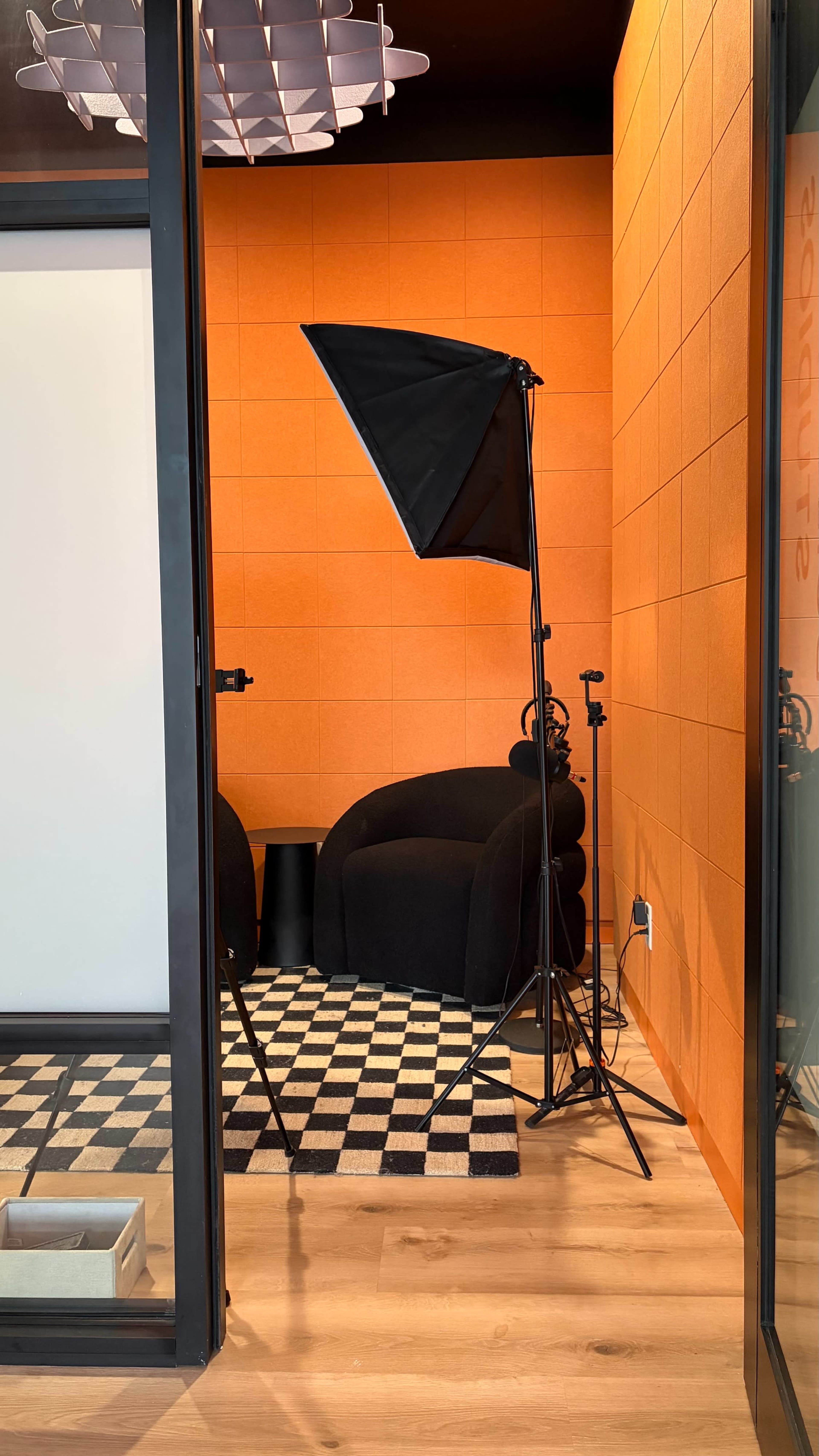 The image depicts a small room with bright orange walls, featuring a black chair, a checkered floor, and lighting equipment set up near a glass entryway.