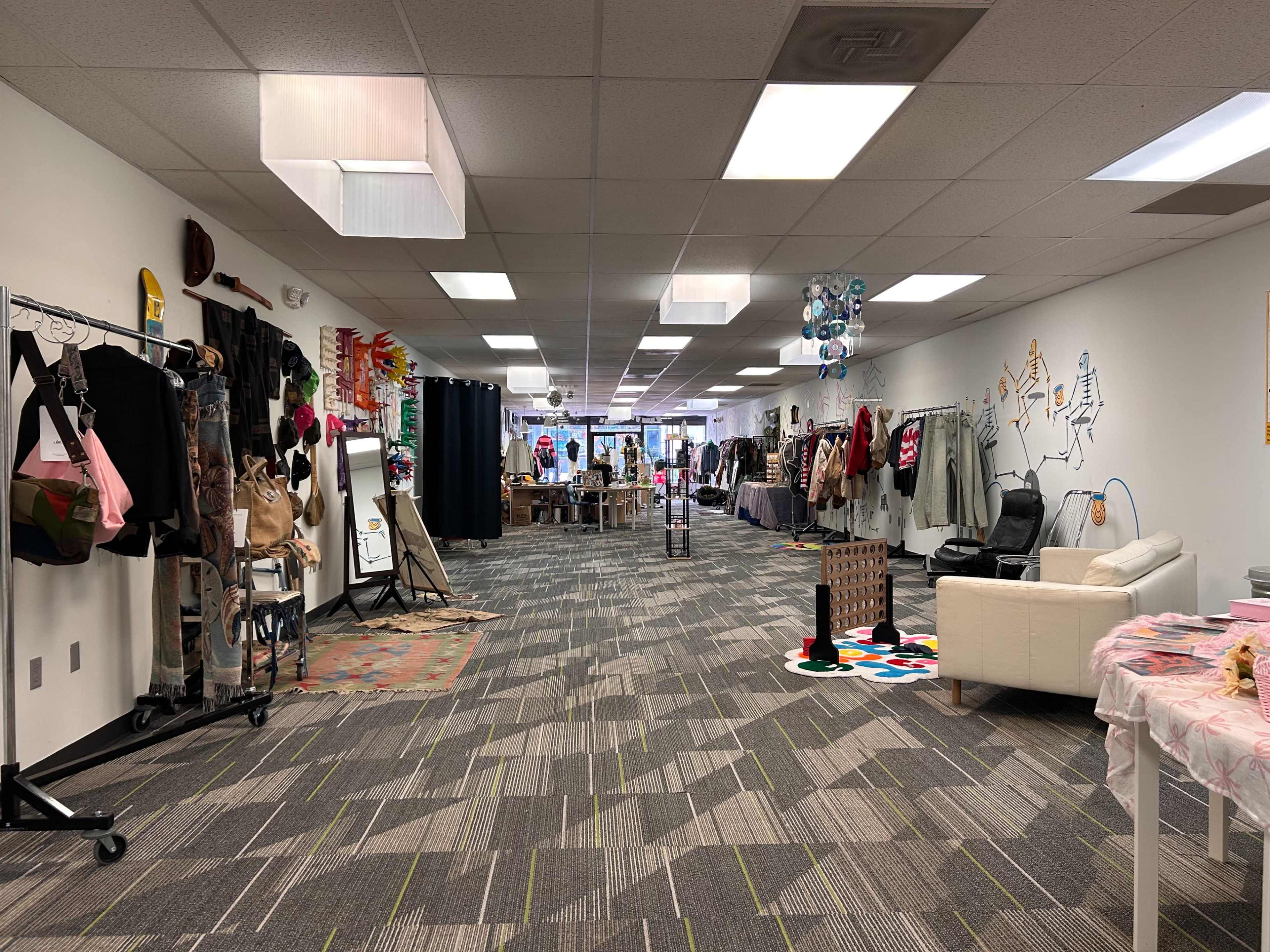 The image shows a spacious indoor area set up as a retail or exhibition space, featuring racks of clothing, tables with various items, and colorful decor along the walls.