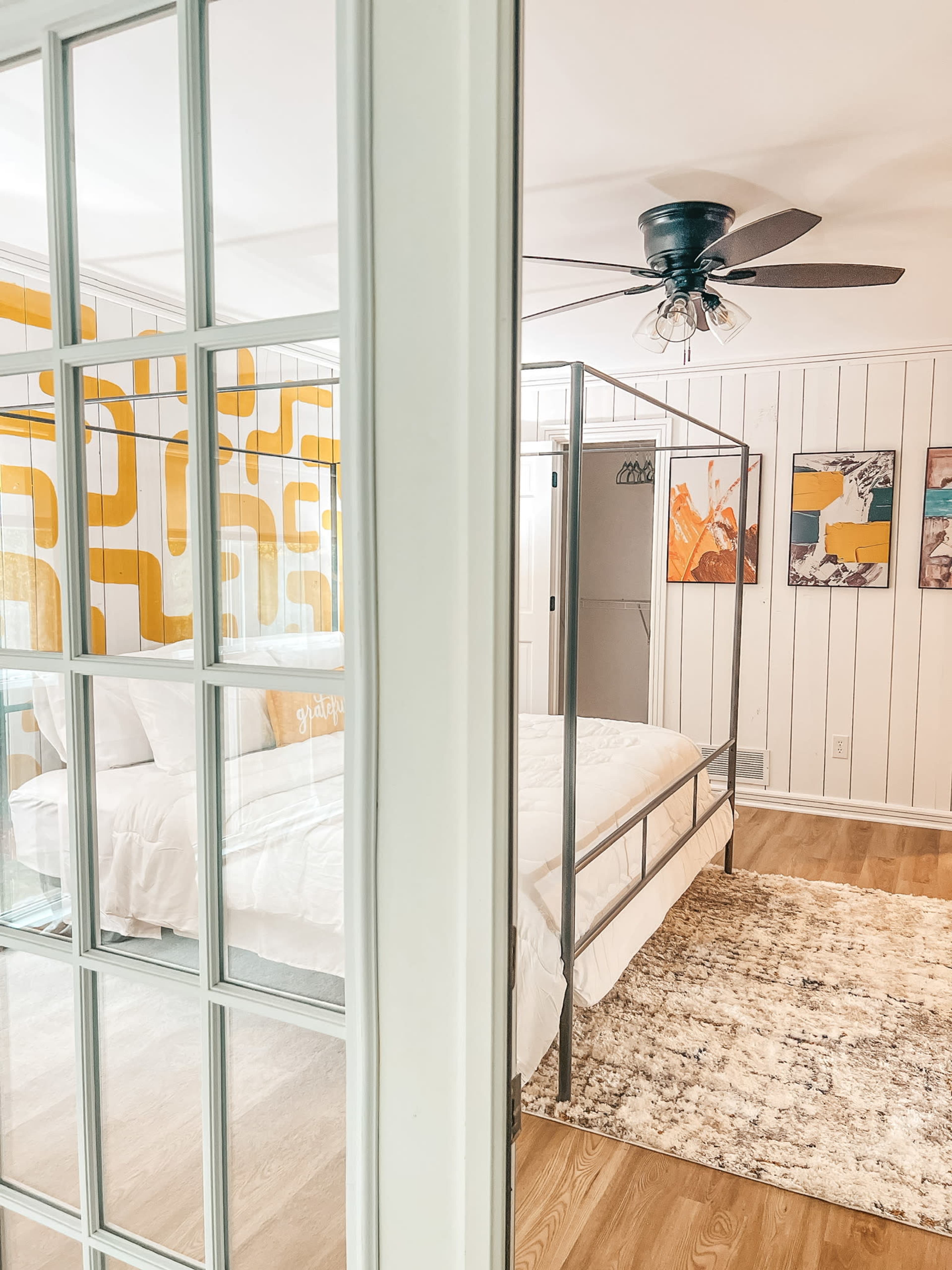 A bright bedroom features a four-poster bed with a white comforter, an accent wall with yellow retro patterns, and framed artwork.