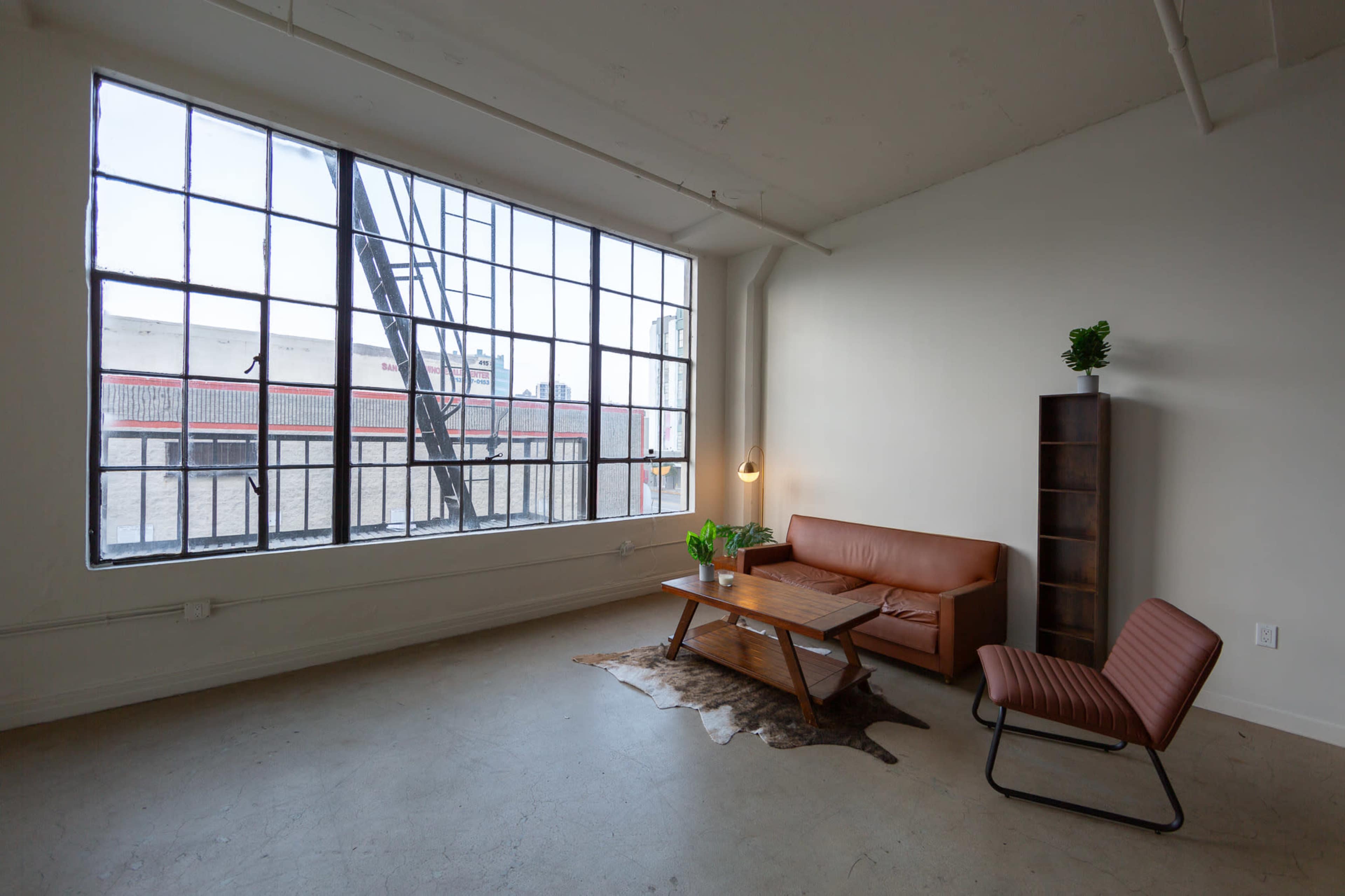 A spacious, minimalistic room with large windows, featuring a brown leather sofa, a wooden coffee table, and a bookshelf against the wall.
