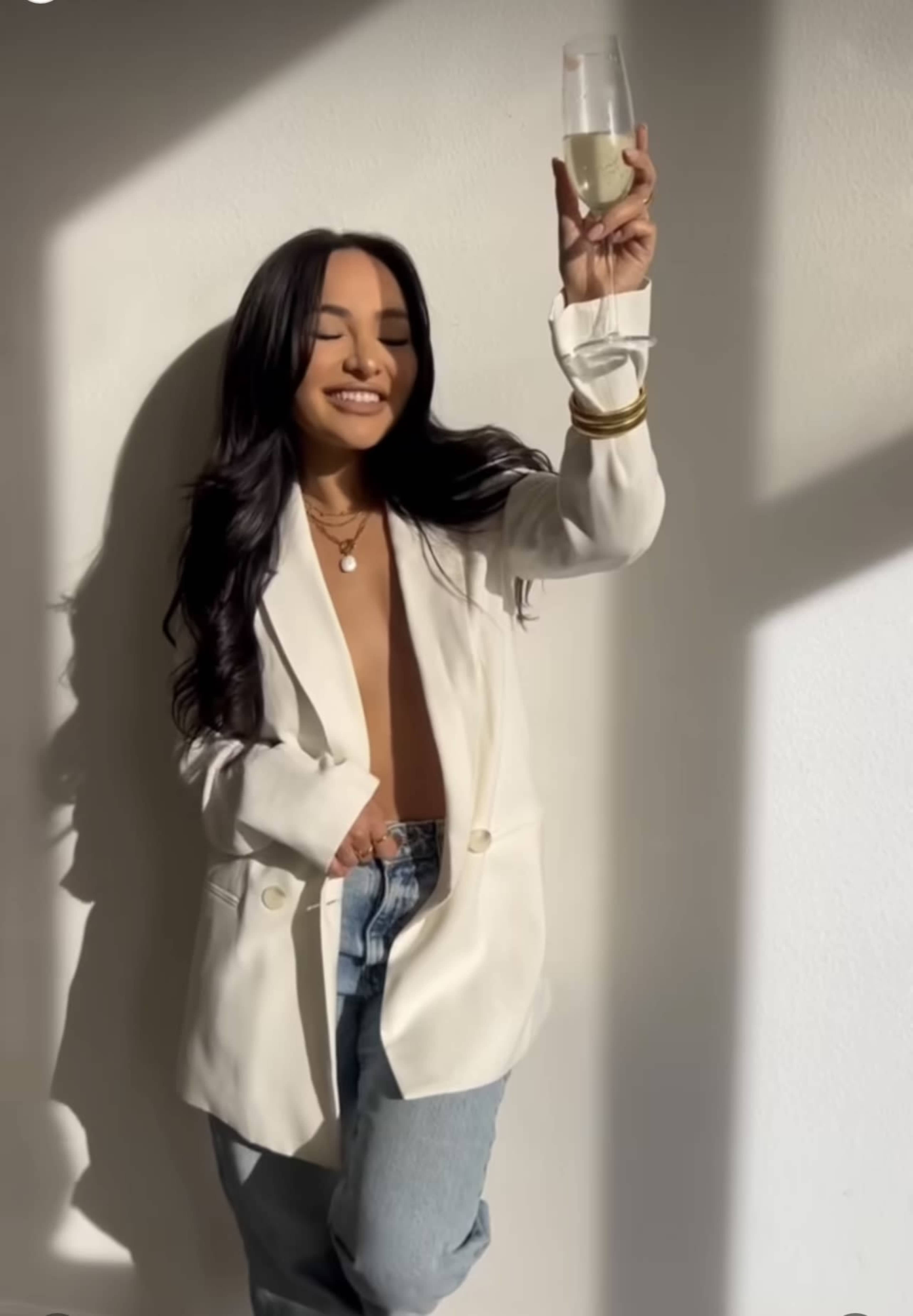 A woman in a white blazer holds a glass of champagne while leaning against a wall.