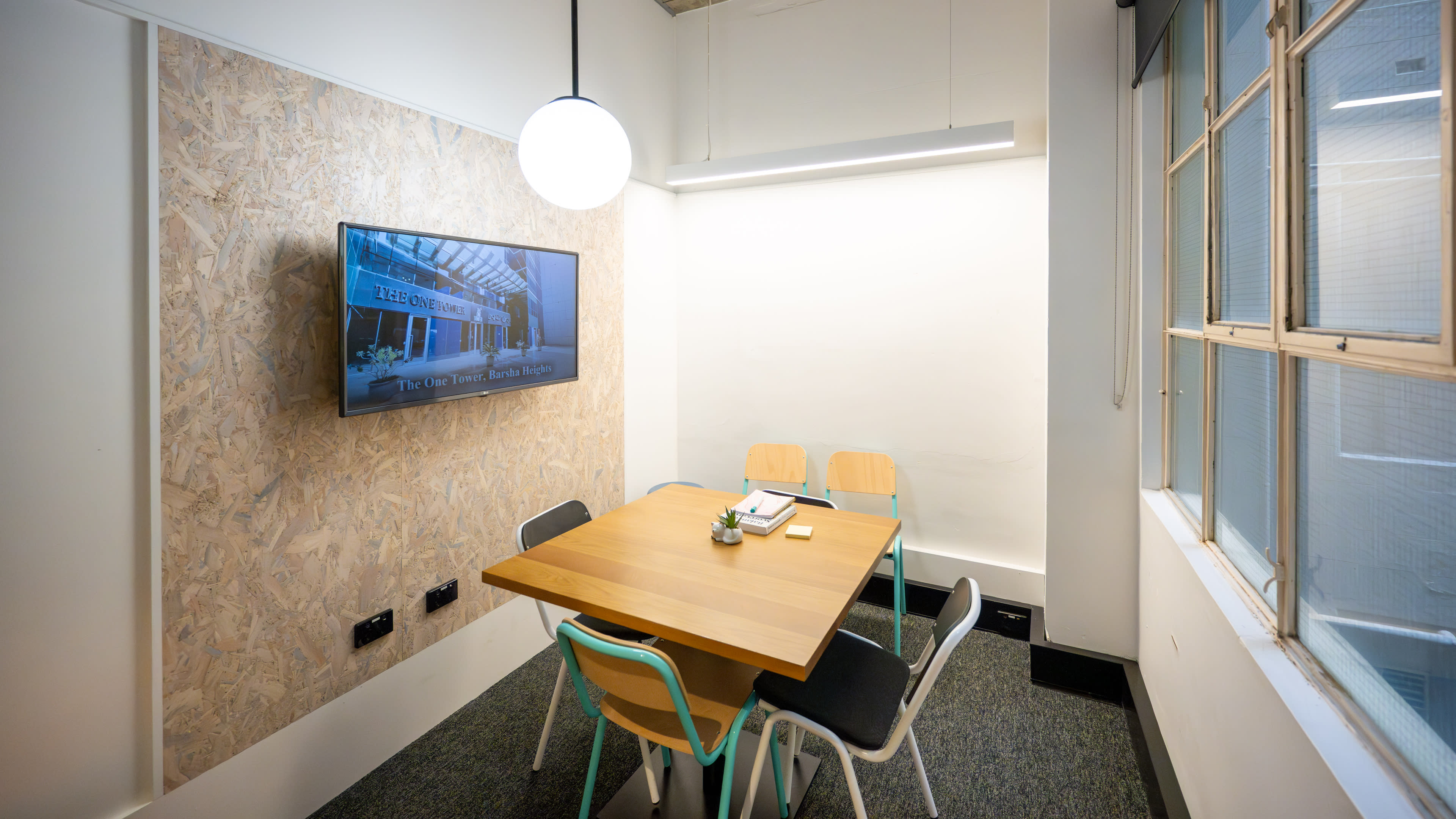 A small meeting room features a wooden table surrounded by four chairs, a wall-mounted television, and large windows that allow natural light.
