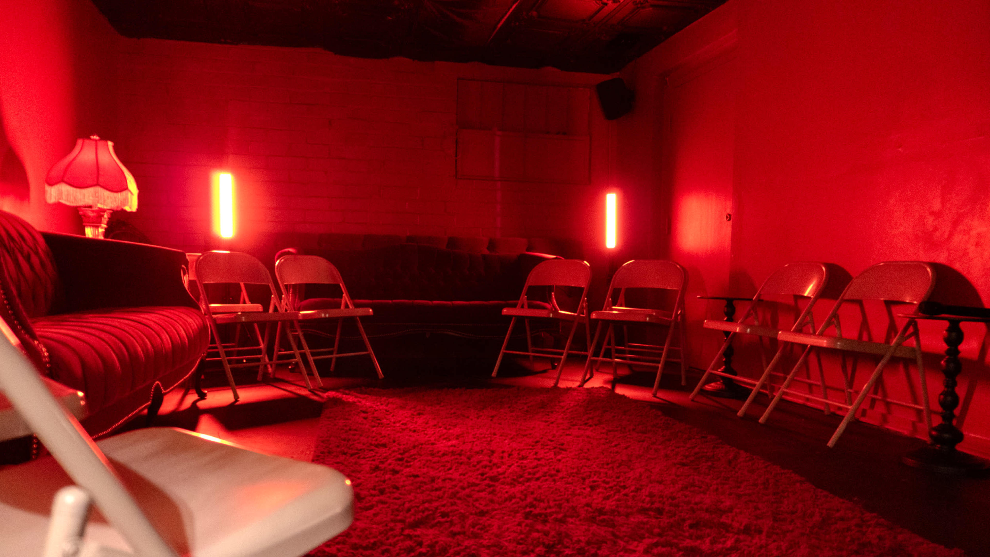The image shows a dimly lit room with red walls, a couch, and several folding chairs arranged in a circle around a central area.