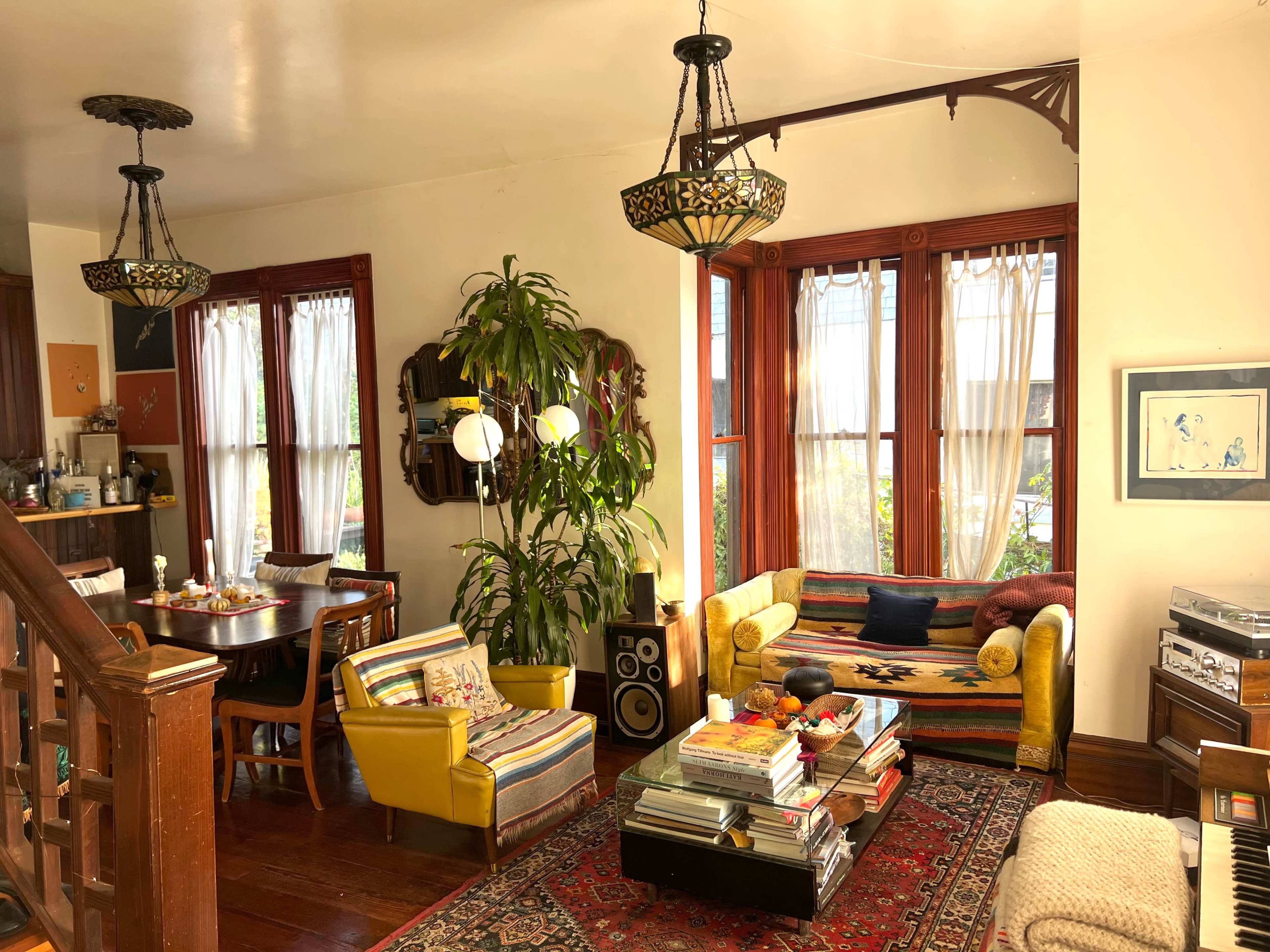 A cozy living room features a mix of furniture, including a colorful couch, yellow chairs, a wooden dining table, and large windows with sheer curtains.