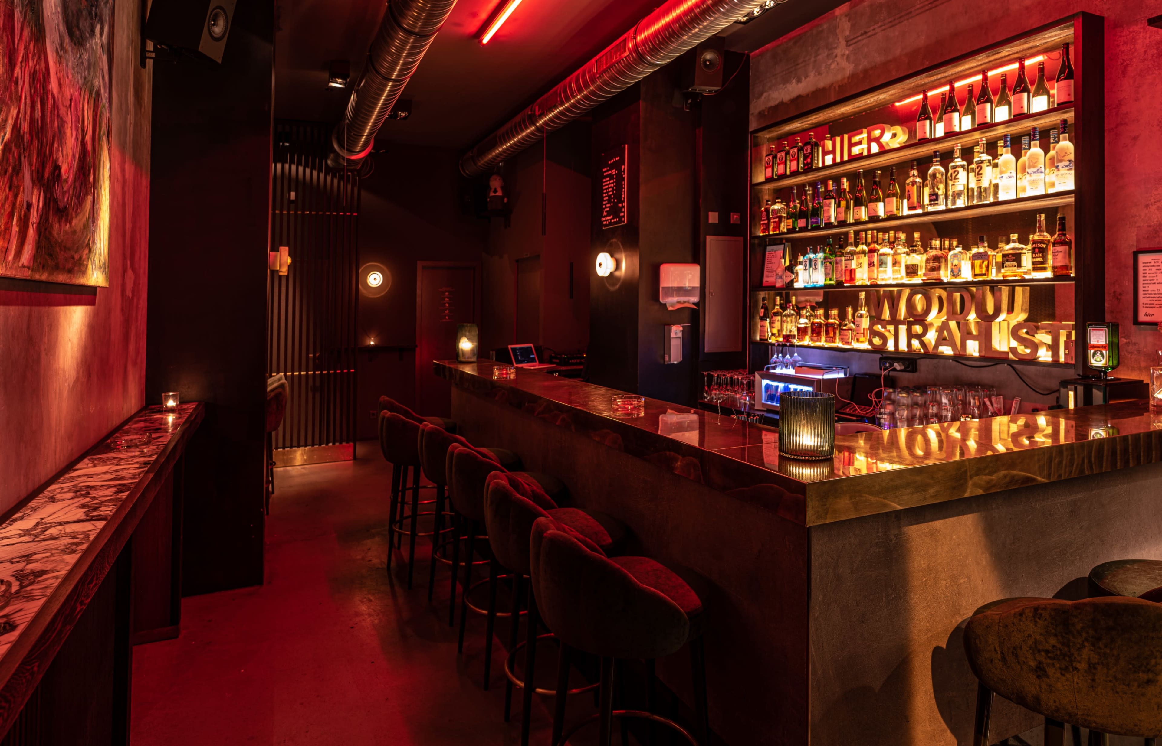 The image shows a dimly lit bar with a long counter, shelves filled with various bottles, and red lighting accents.