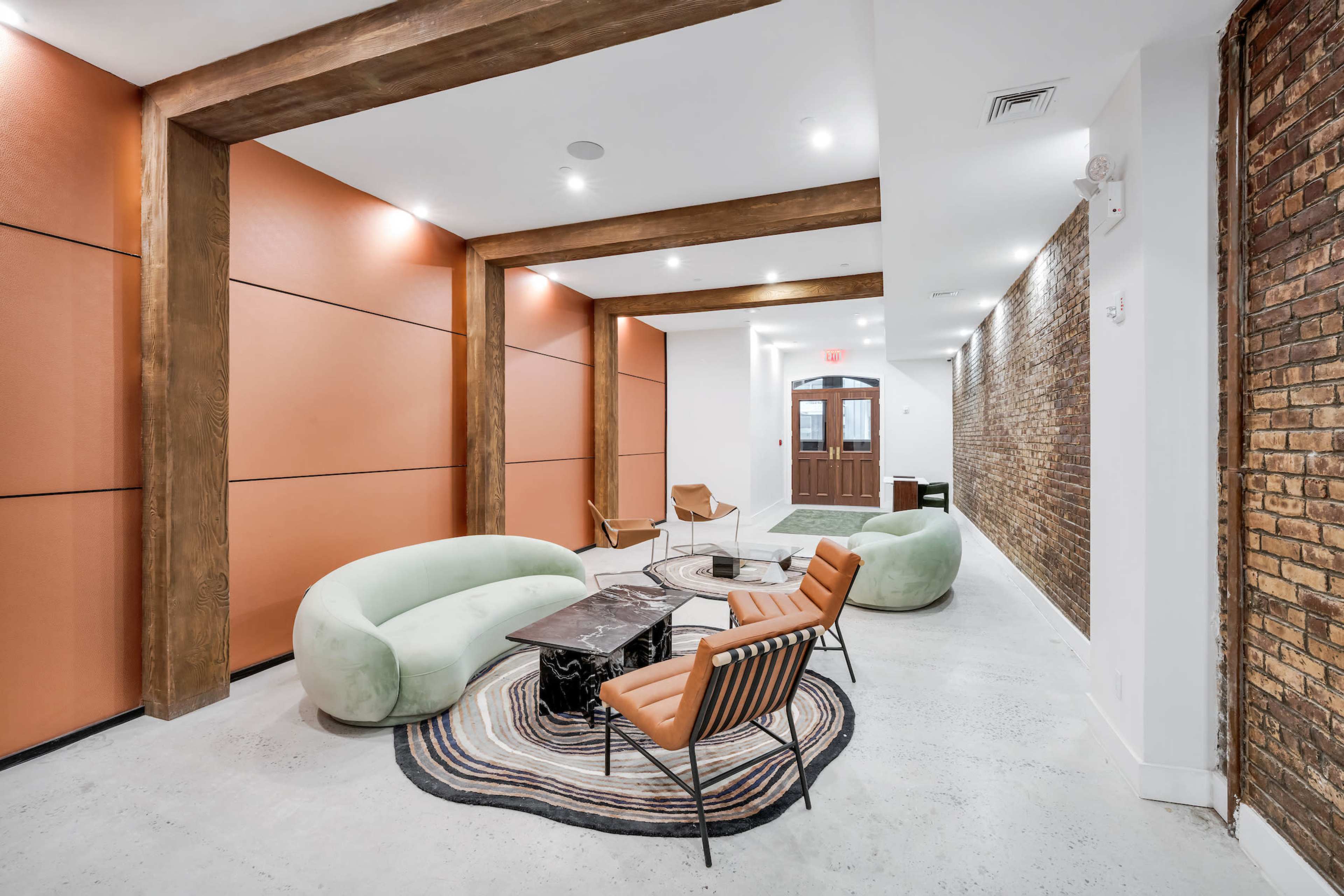 The image shows a modern interior lounge area with curved green sofas, a wooden coffee table, and a mixture of brick and orange walls.