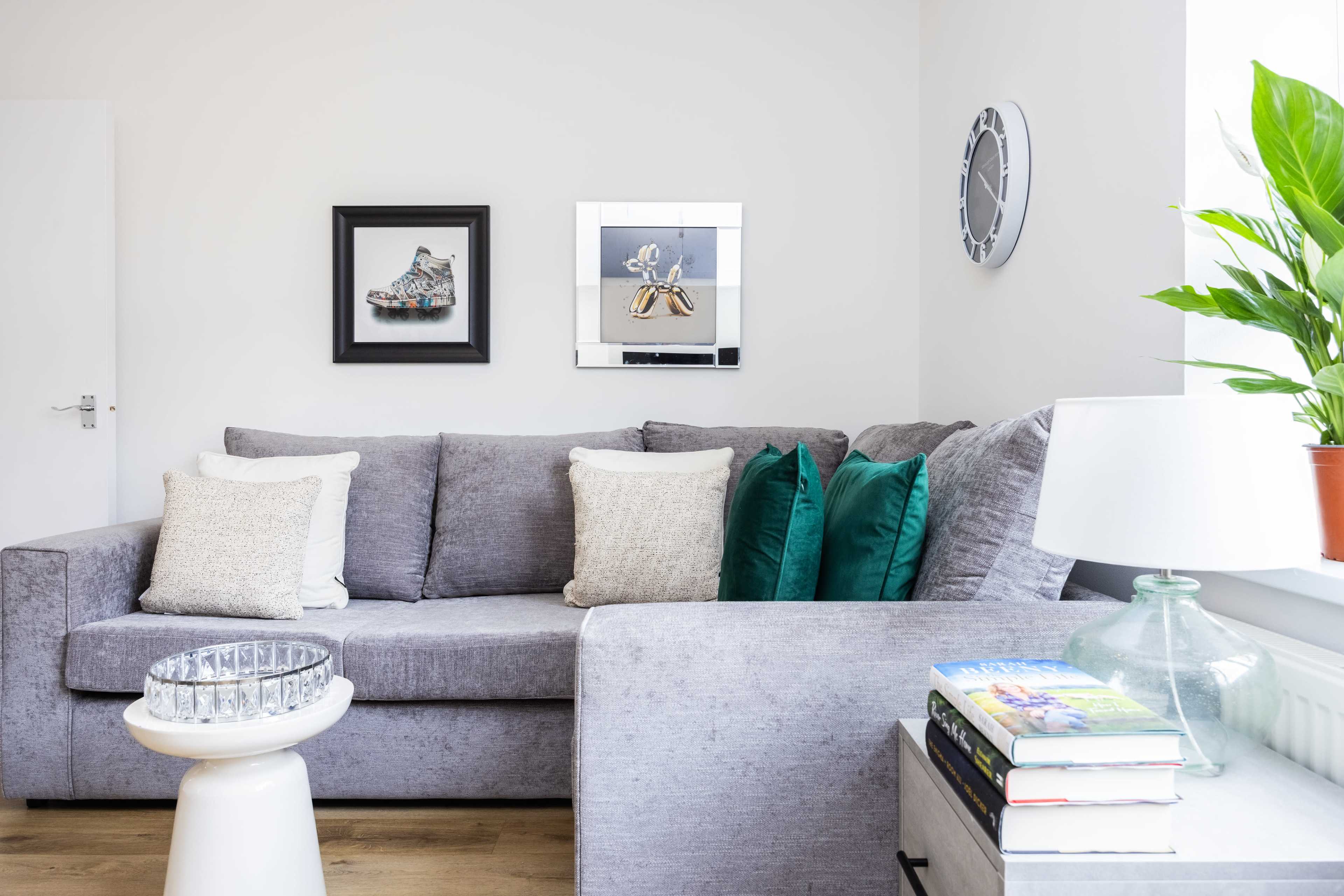 A gray sectional sofa with decorative cushions is placed in a well-lit room featuring artwork on the walls and a lamp on a side table.