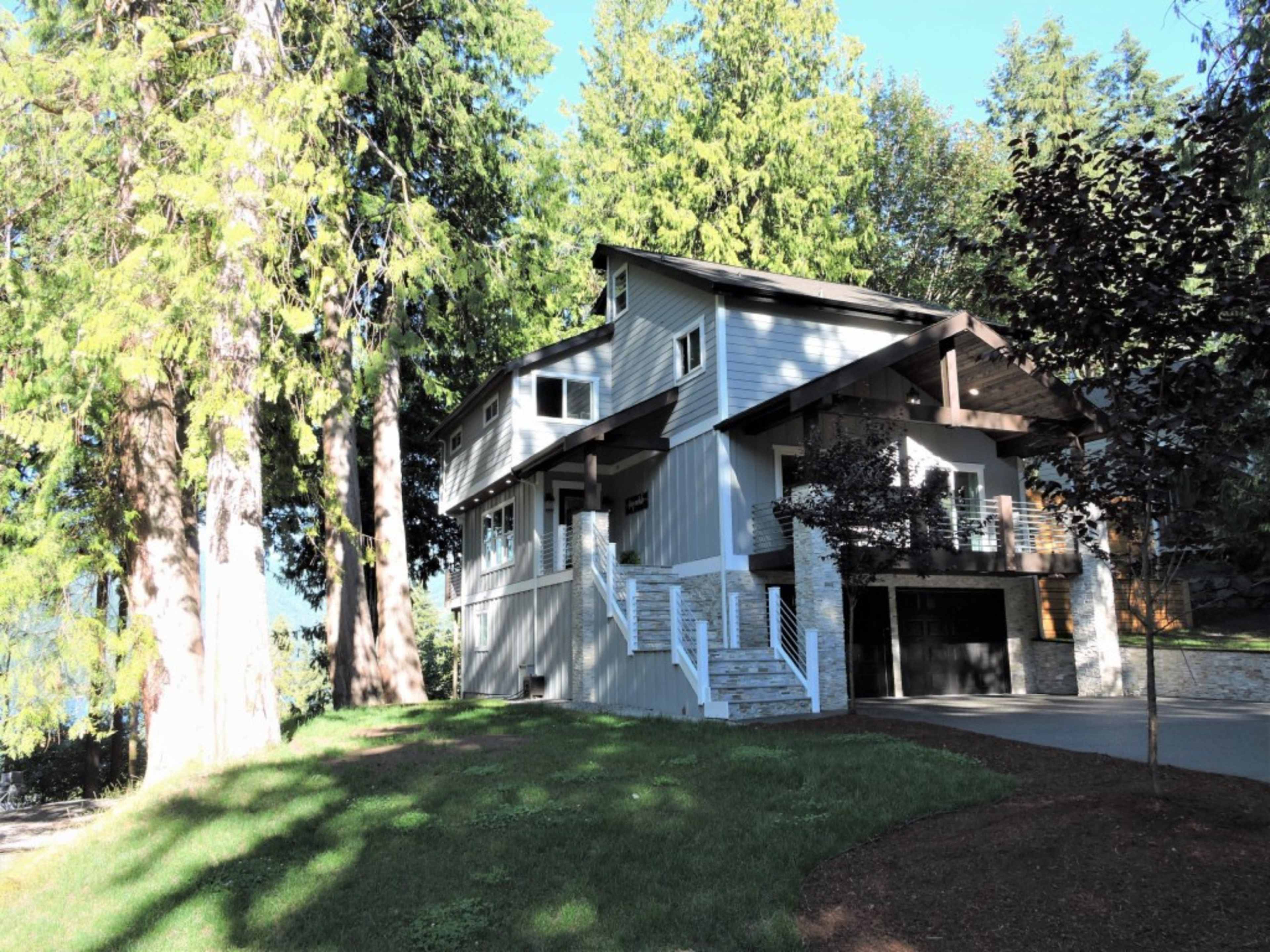 A two-story house with a garage and a front staircase is surrounded by tall trees and lush greenery.