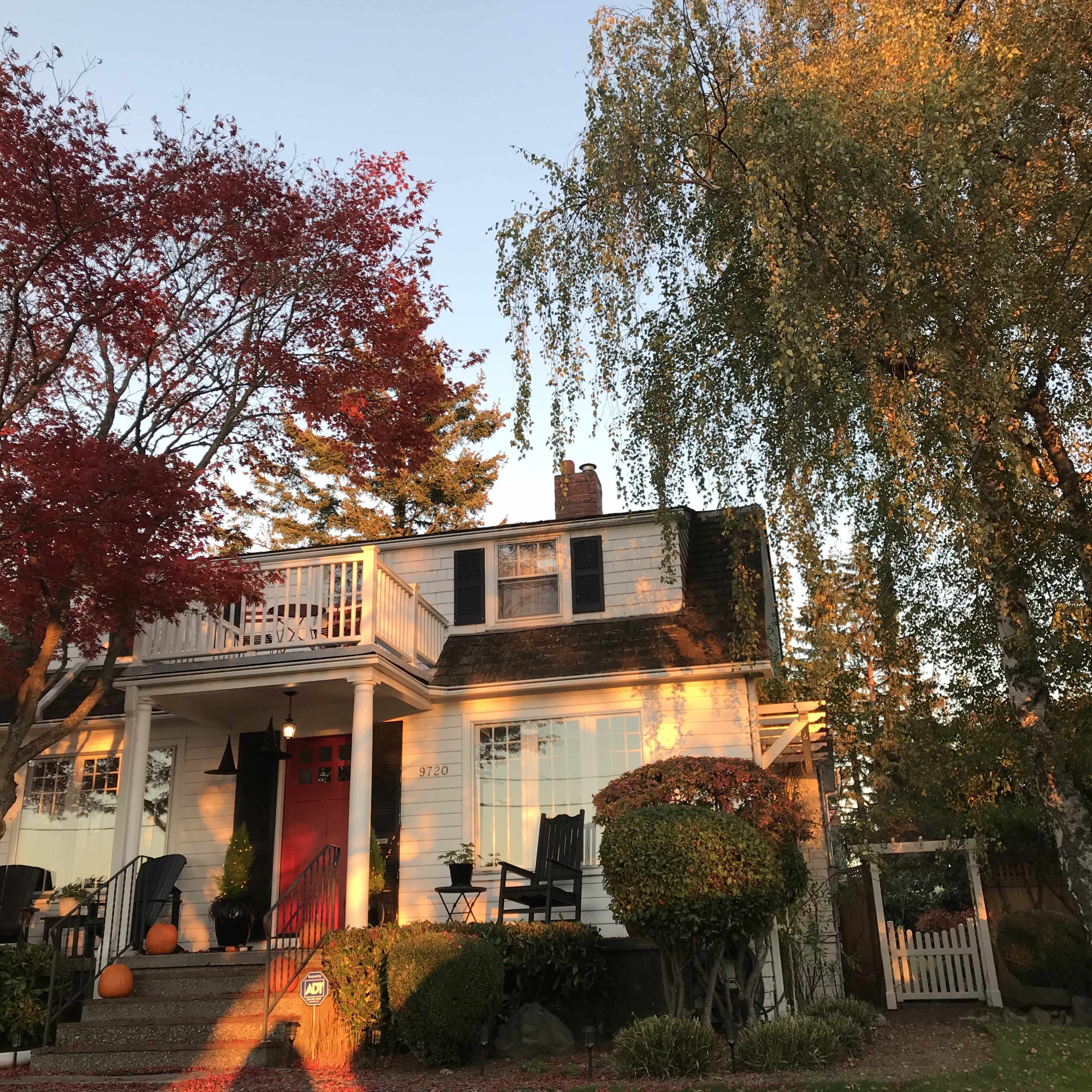 The house has a red front door, a balcony, and is surrounded by trees with colorful foliage.