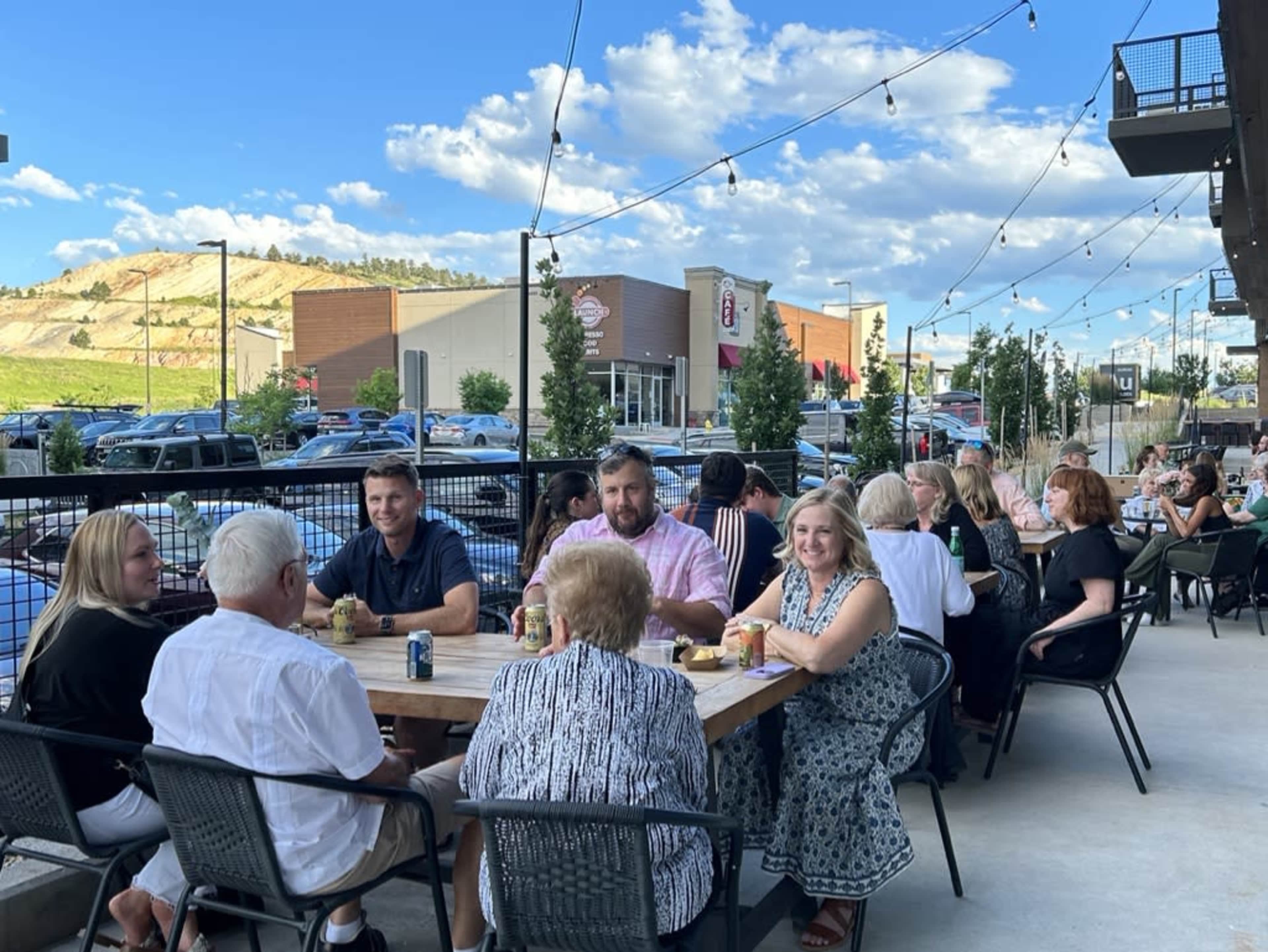 A group of people sits around a large table on a patio, enjoying drinks and food, with a backdrop of a parking lot and hills under a blue sky.