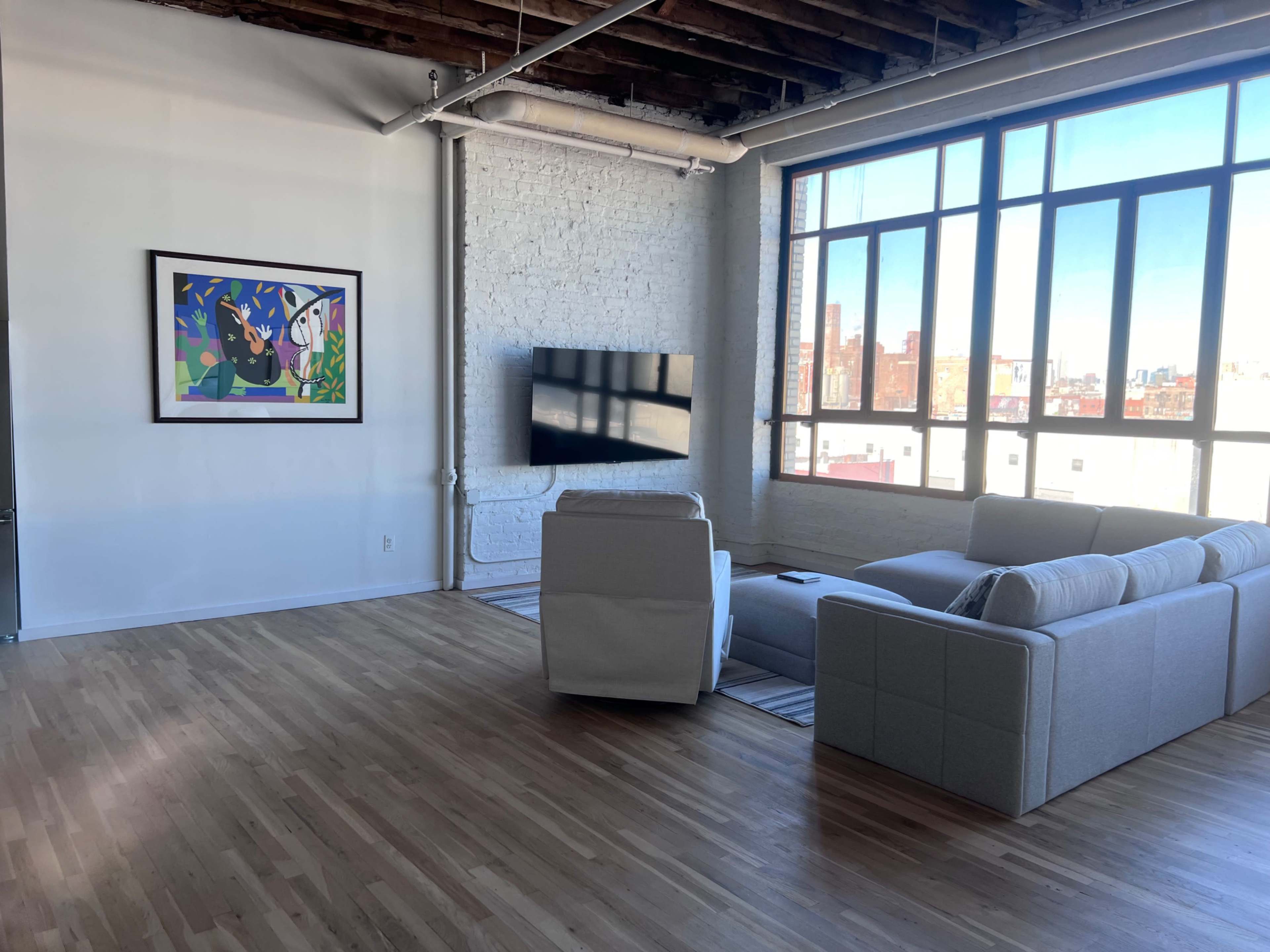 A modern living room features a large window providing city views, a flat-screen TV mounted on the wall, and a sectional sofa arranged on wooden flooring.