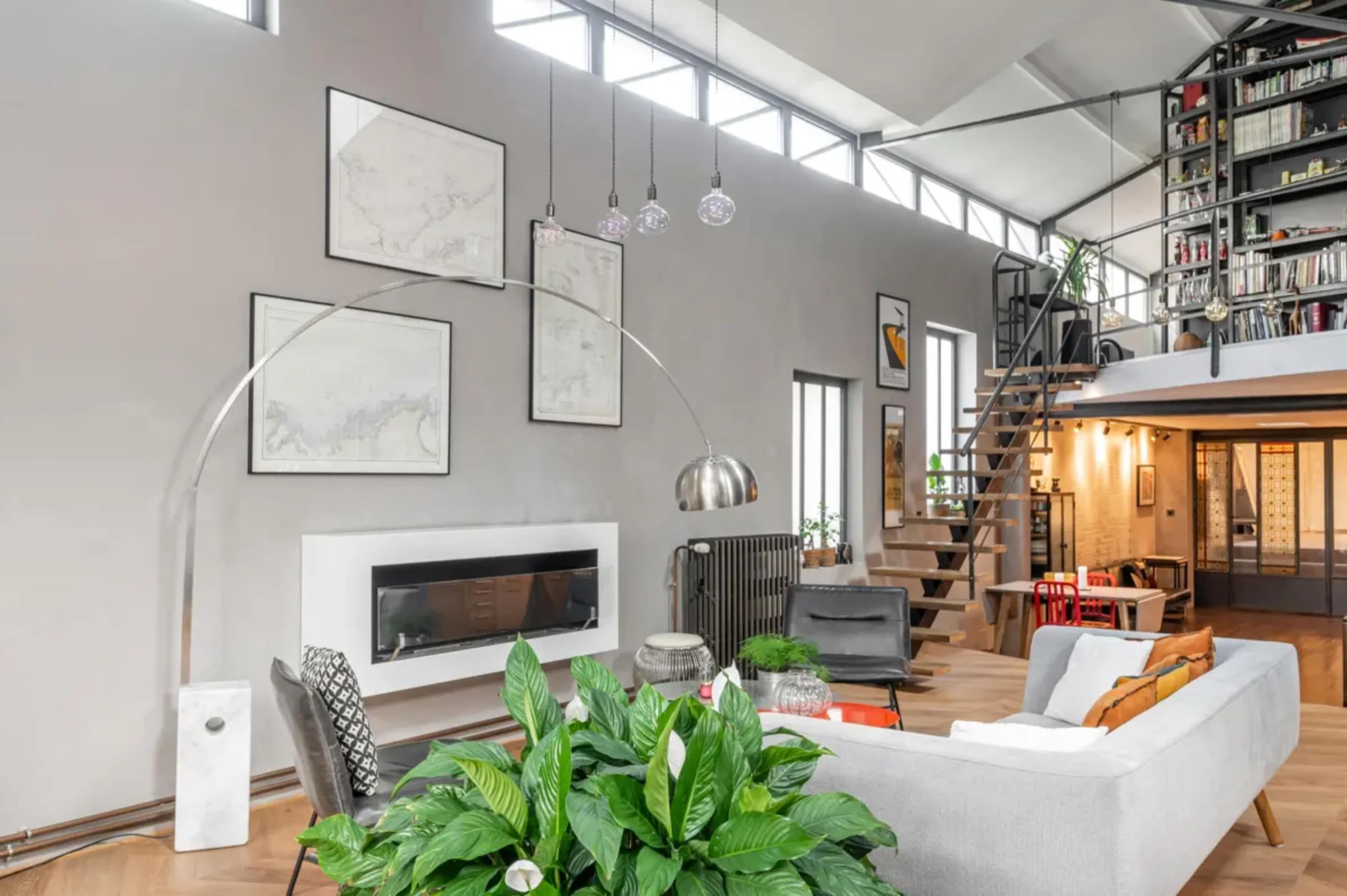 A modern living room featuring a gray sectional sofa, a contemporary arc floor lamp, a wall-mounted fireplace, and a staircase that leads to a loft area.