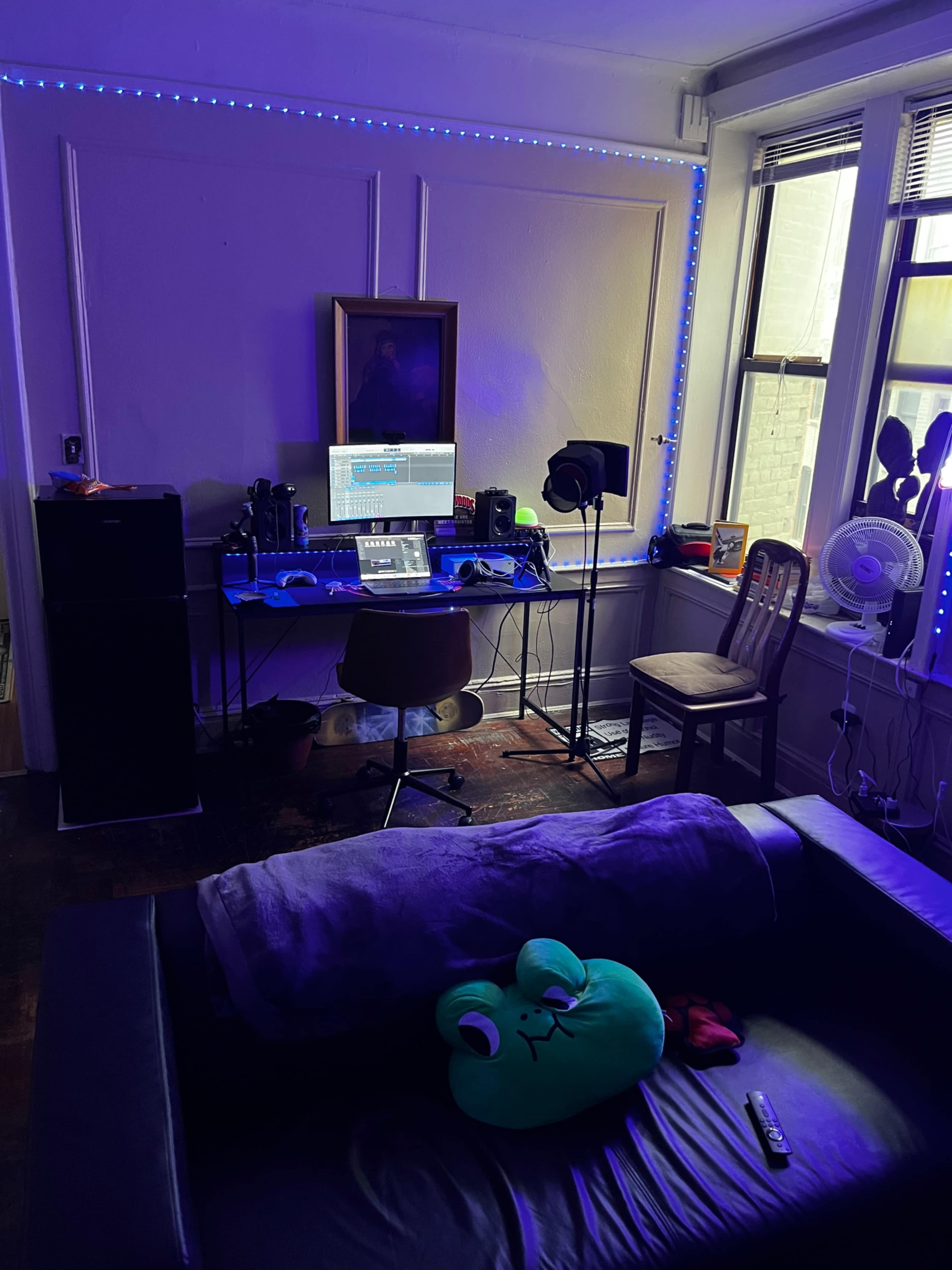 A room with a dark couch, a desk equipped with a computer and music production equipment, and ambient LED lights illuminating the walls.