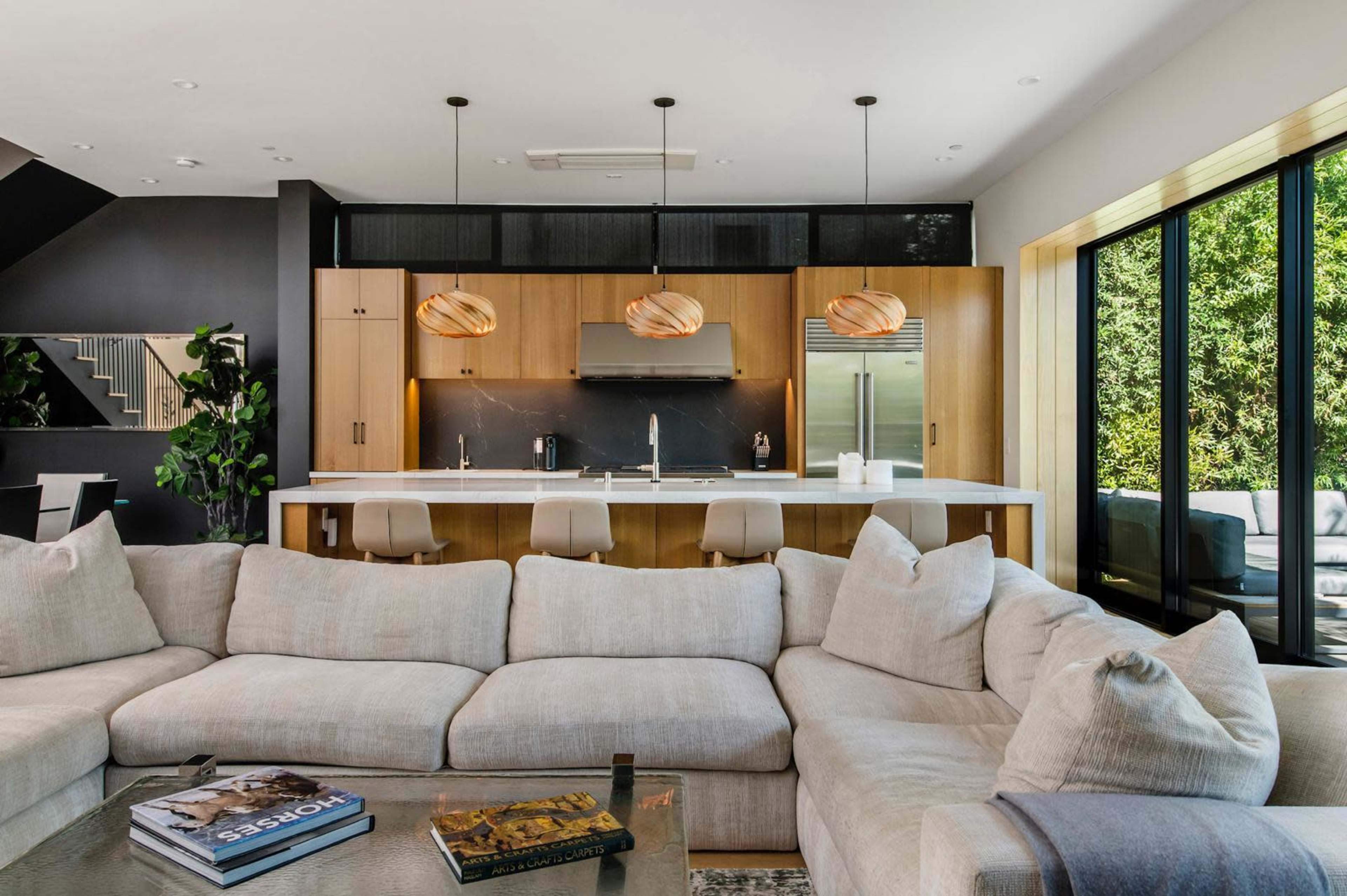 The image shows a modern living room with a large, light-colored sectional sofa, a coffee table with books, and a kitchen area featuring wooden cabinetry and pendant lighting.