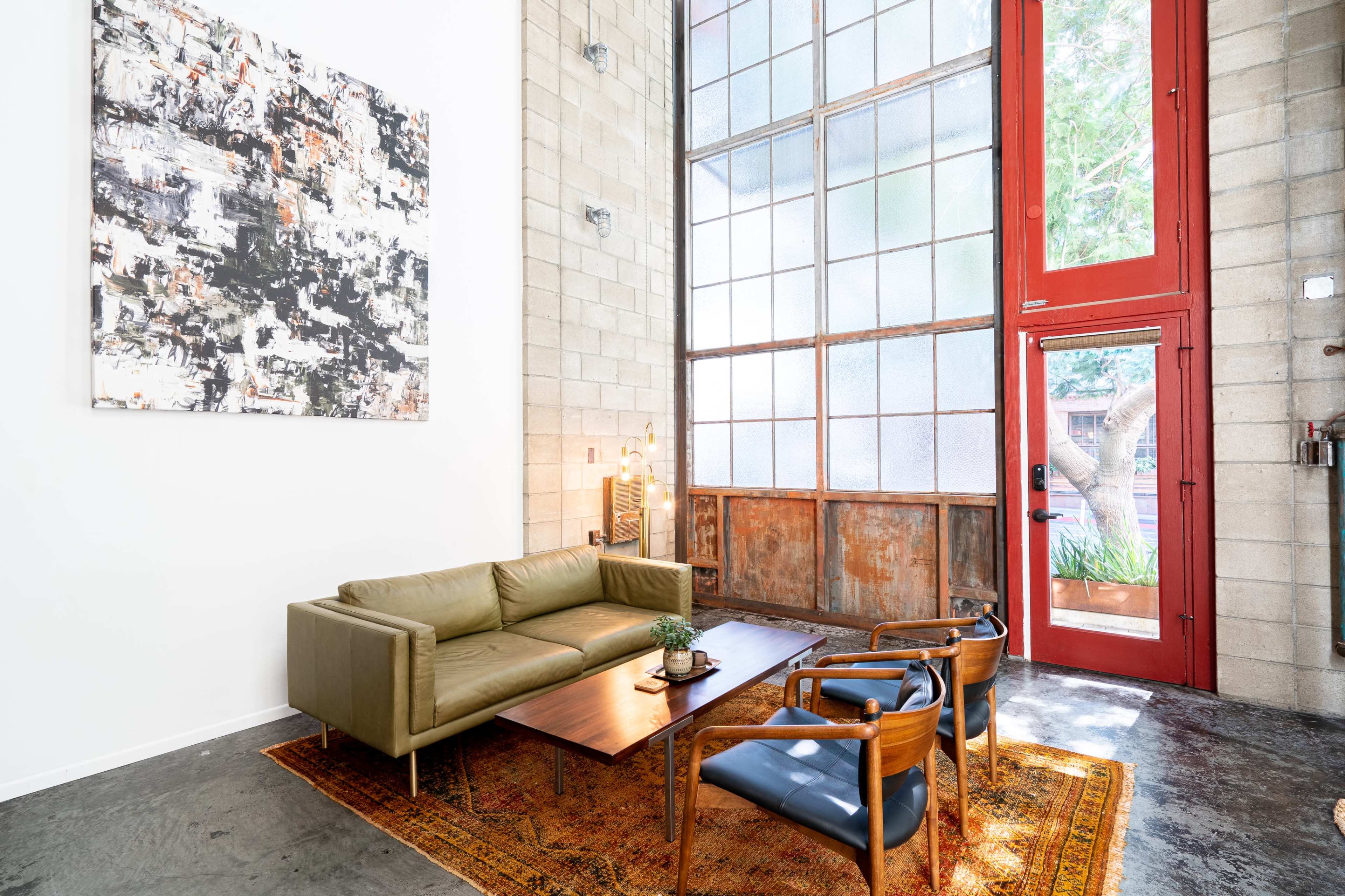 The image shows a modern living area with a green sofa, two wooden chairs, a coffee table, and large windows, all set against a backdrop of textured concrete walls.