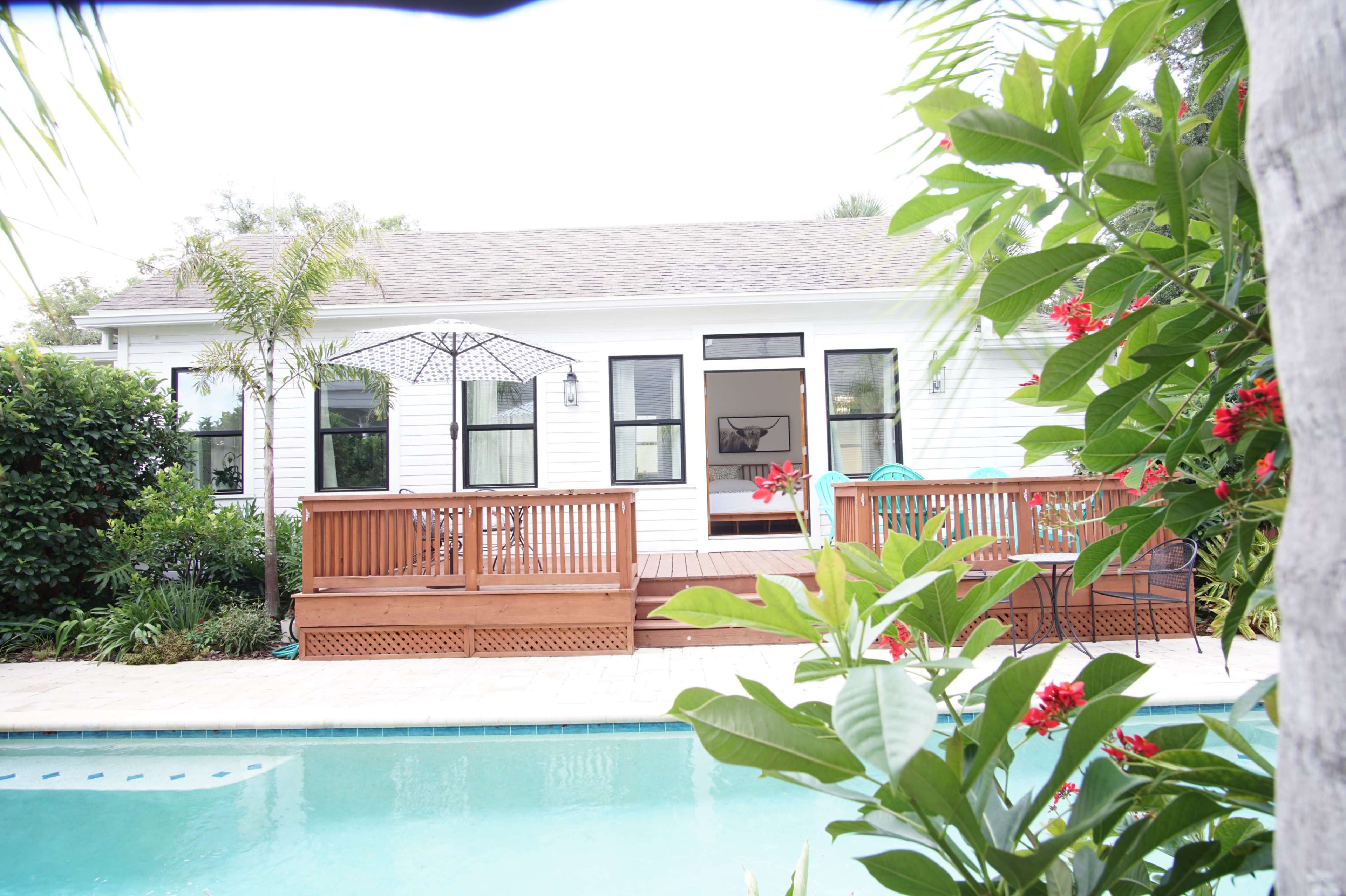 A wooden deck leads to a white house with large windows, surrounded by lush greenery and a swimming pool in the foreground.