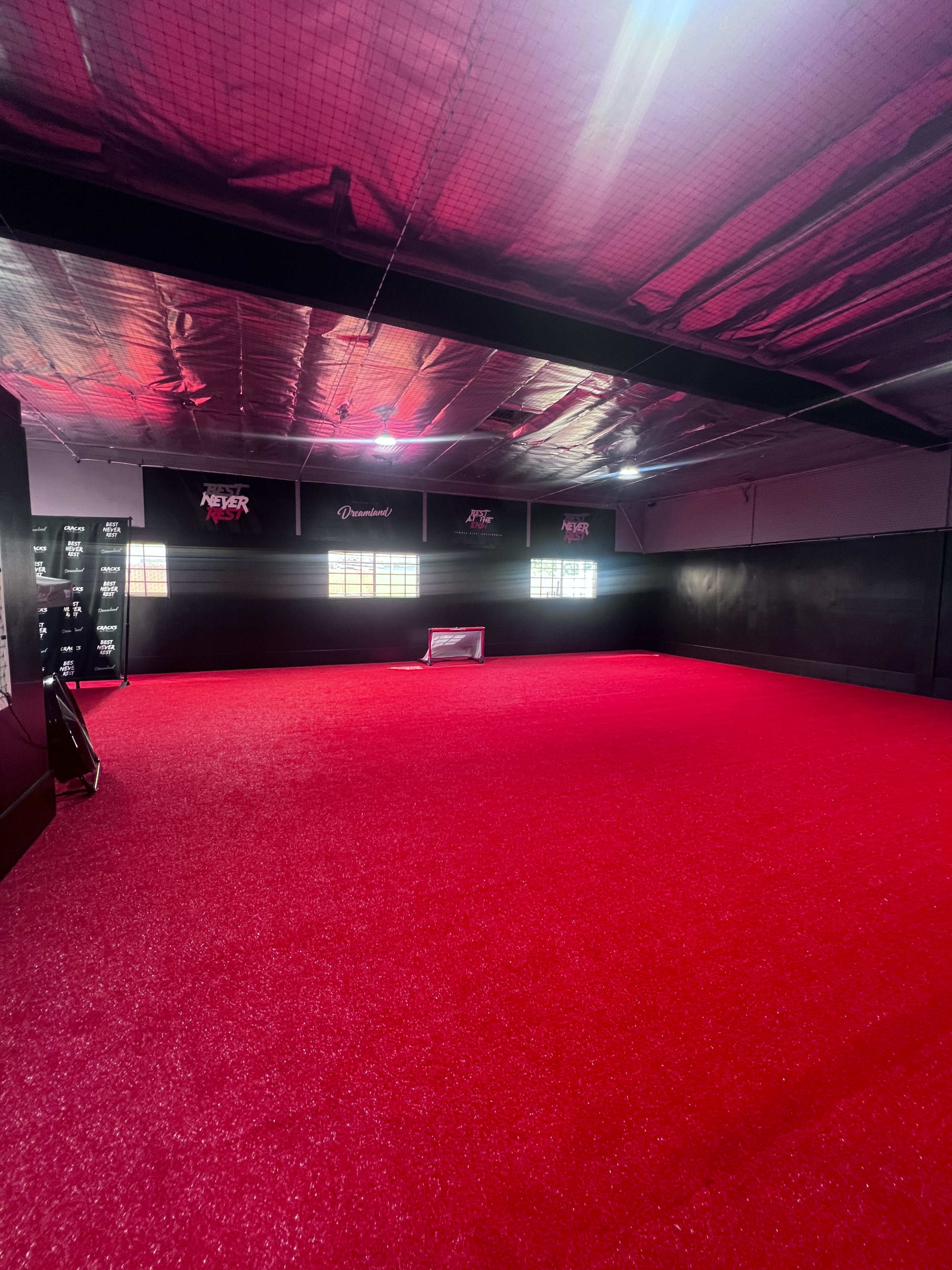 The image shows a spacious indoor sports facility with a bright red synthetic surface, black walls, and overhead lights.