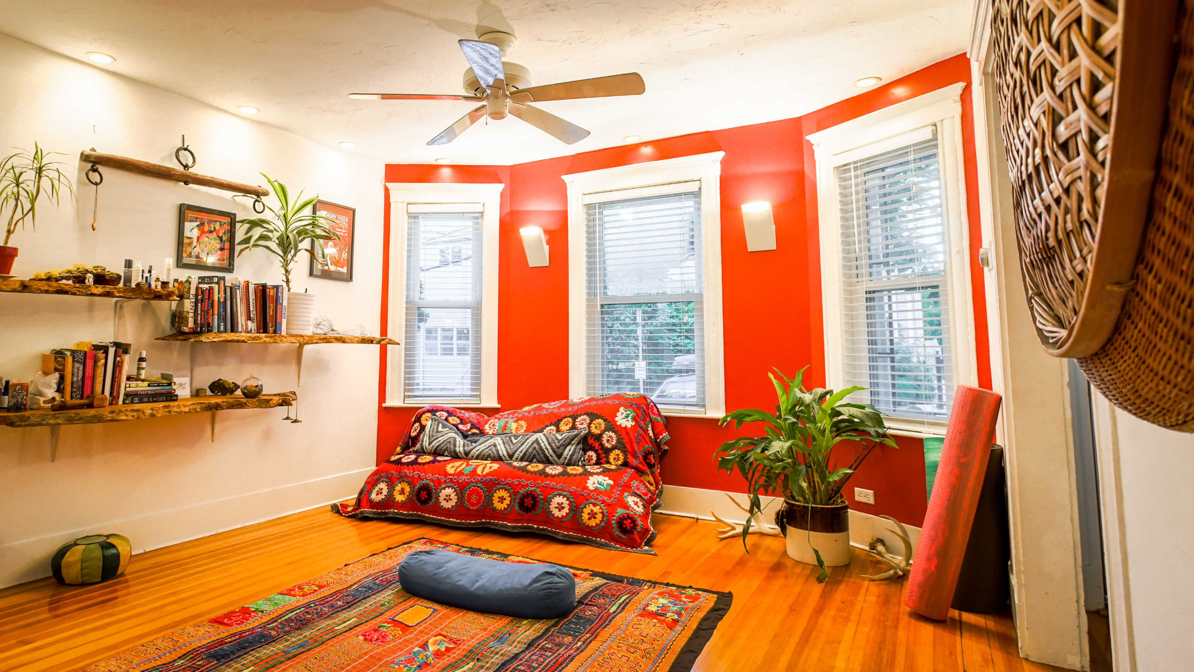 The image shows a cozy room with a red accent wall, a bookshelf, a patterned couch, and a small indoor plant in the corner.