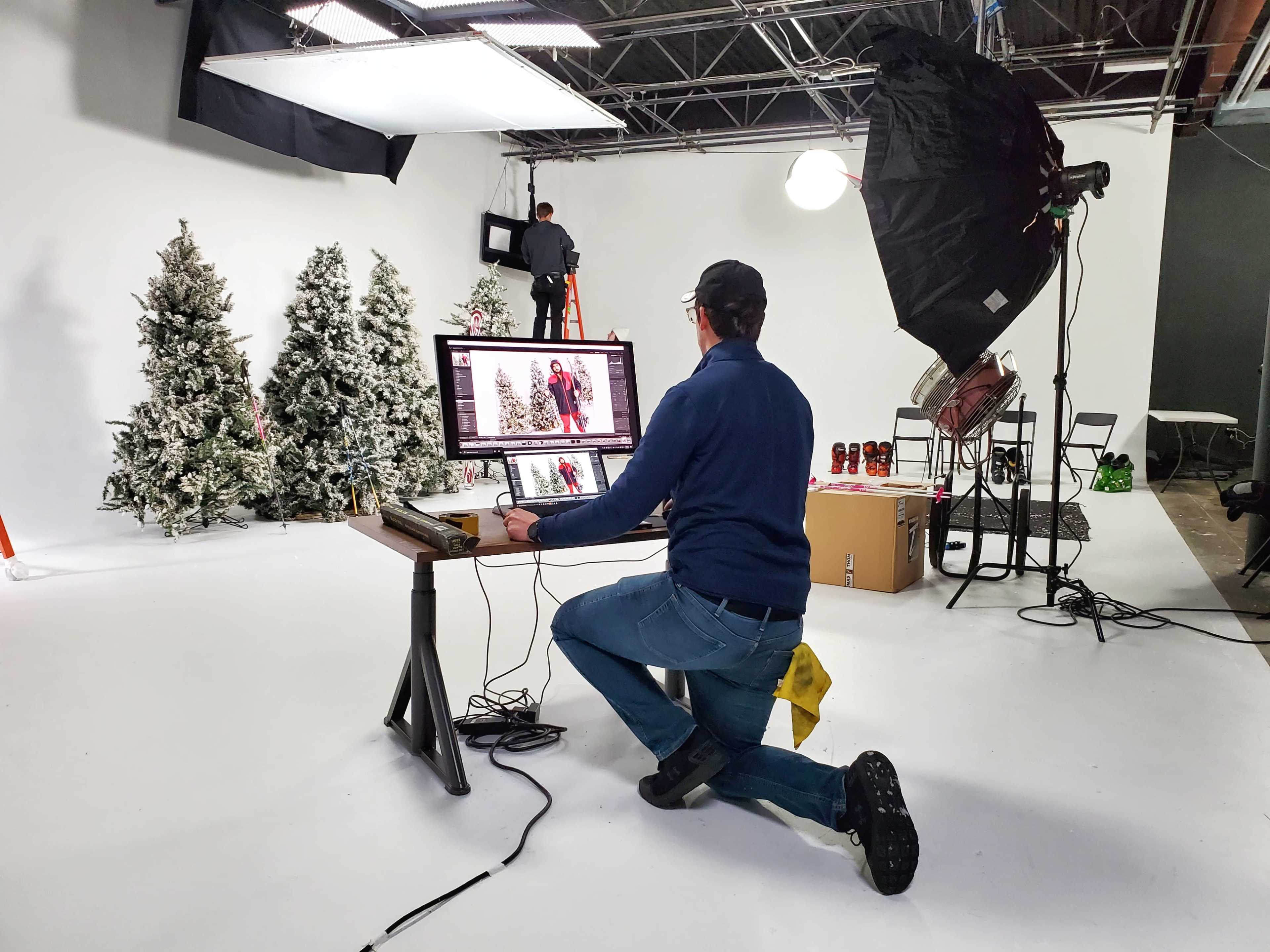 A photographer edits images on a computer while kneeling in a studio decorated with artificial Christmas trees and various equipment.
