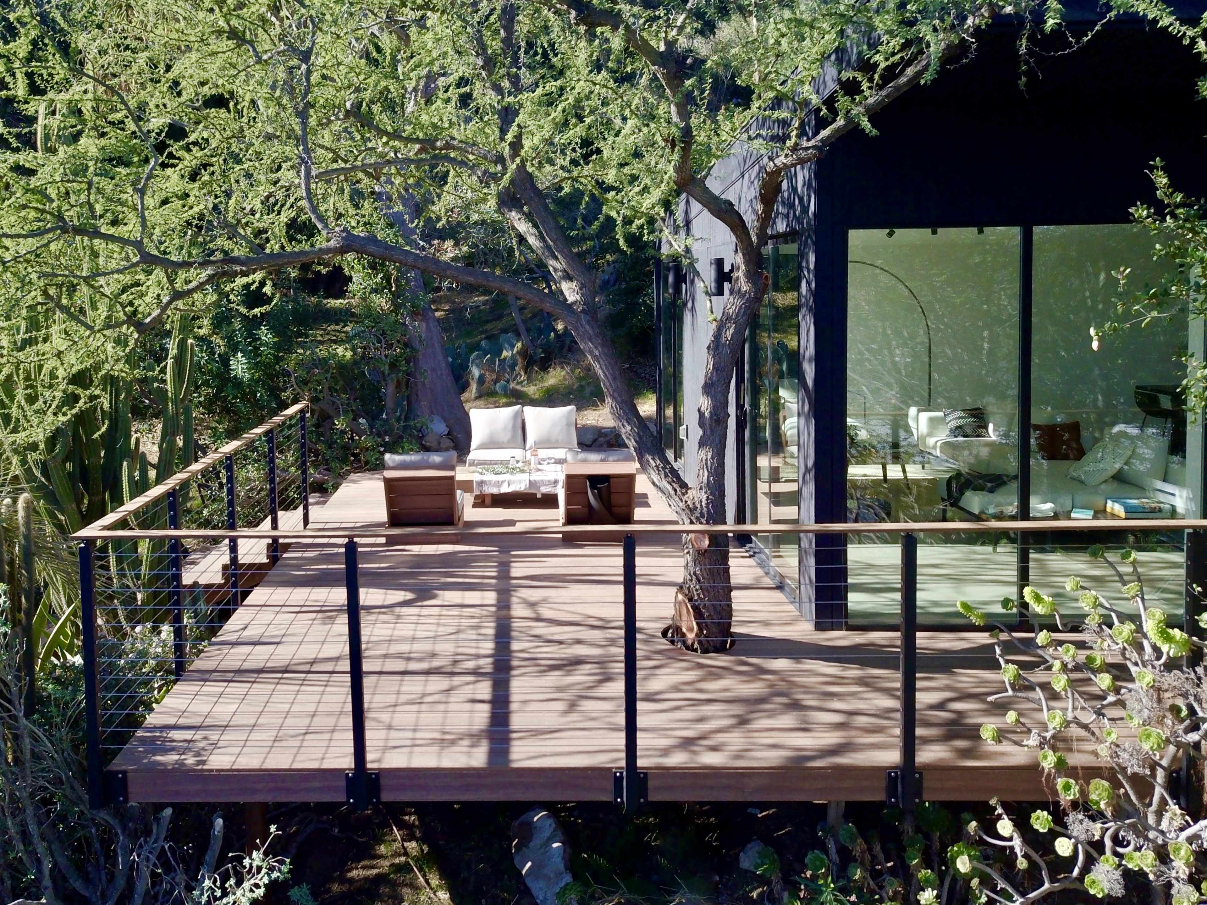 A modern wooden deck features outdoor seating beside a house with large glass windows, surrounded by greenery and a prominent tree.