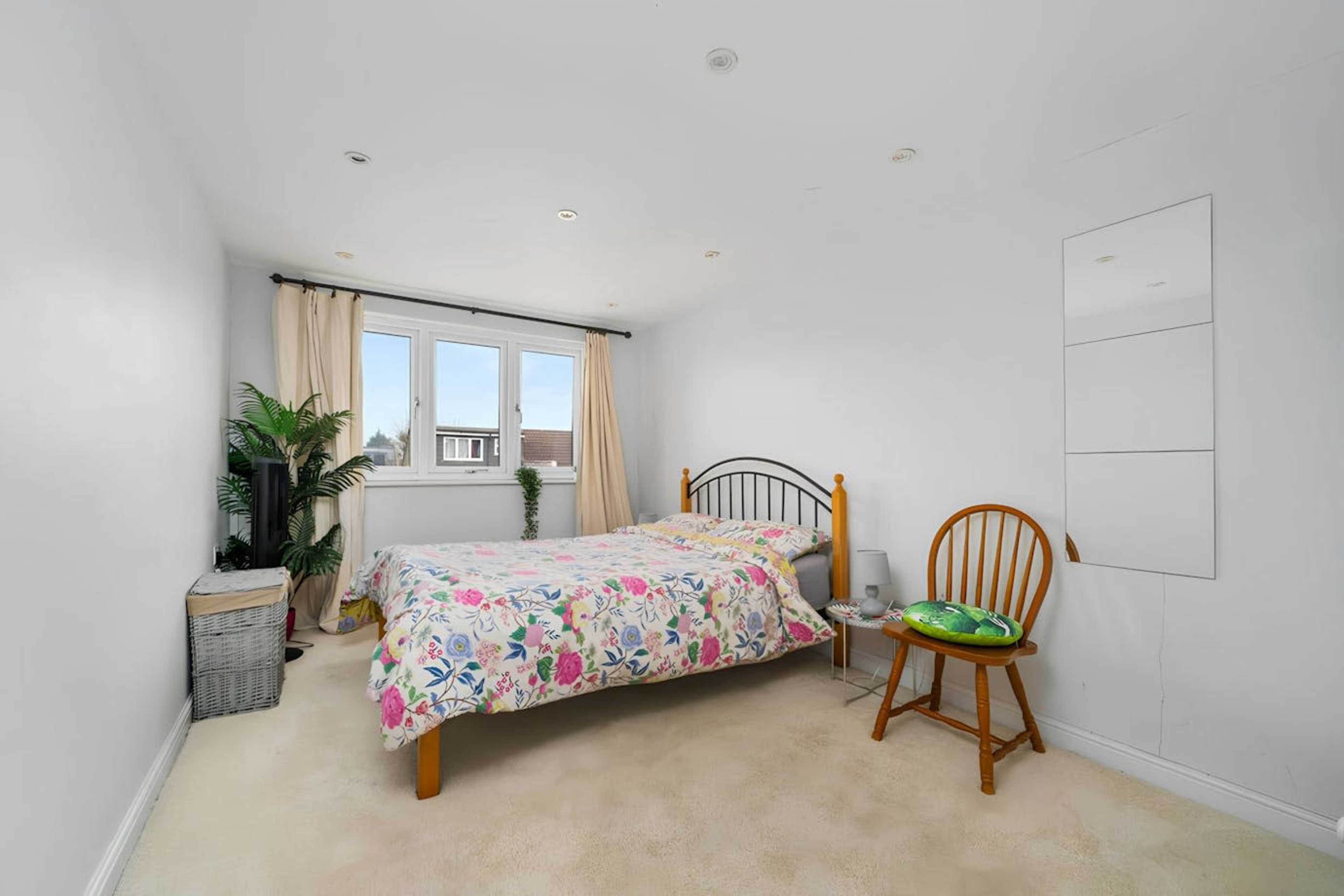 A simple bedroom features a bed with a floral blanket, a wooden chair, and a small storage basket, with a window allowing natural light to enter the space.