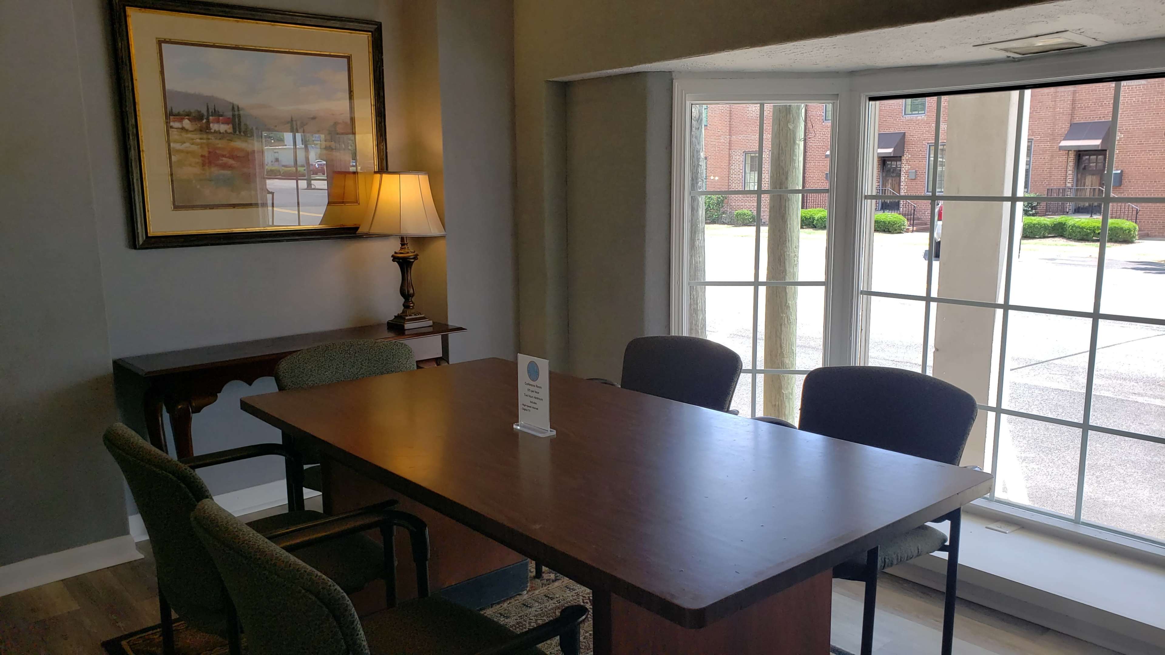 A small meeting room with a table surrounded by four chairs, a lamp on a side table, and large windows allowing natural light to enter.