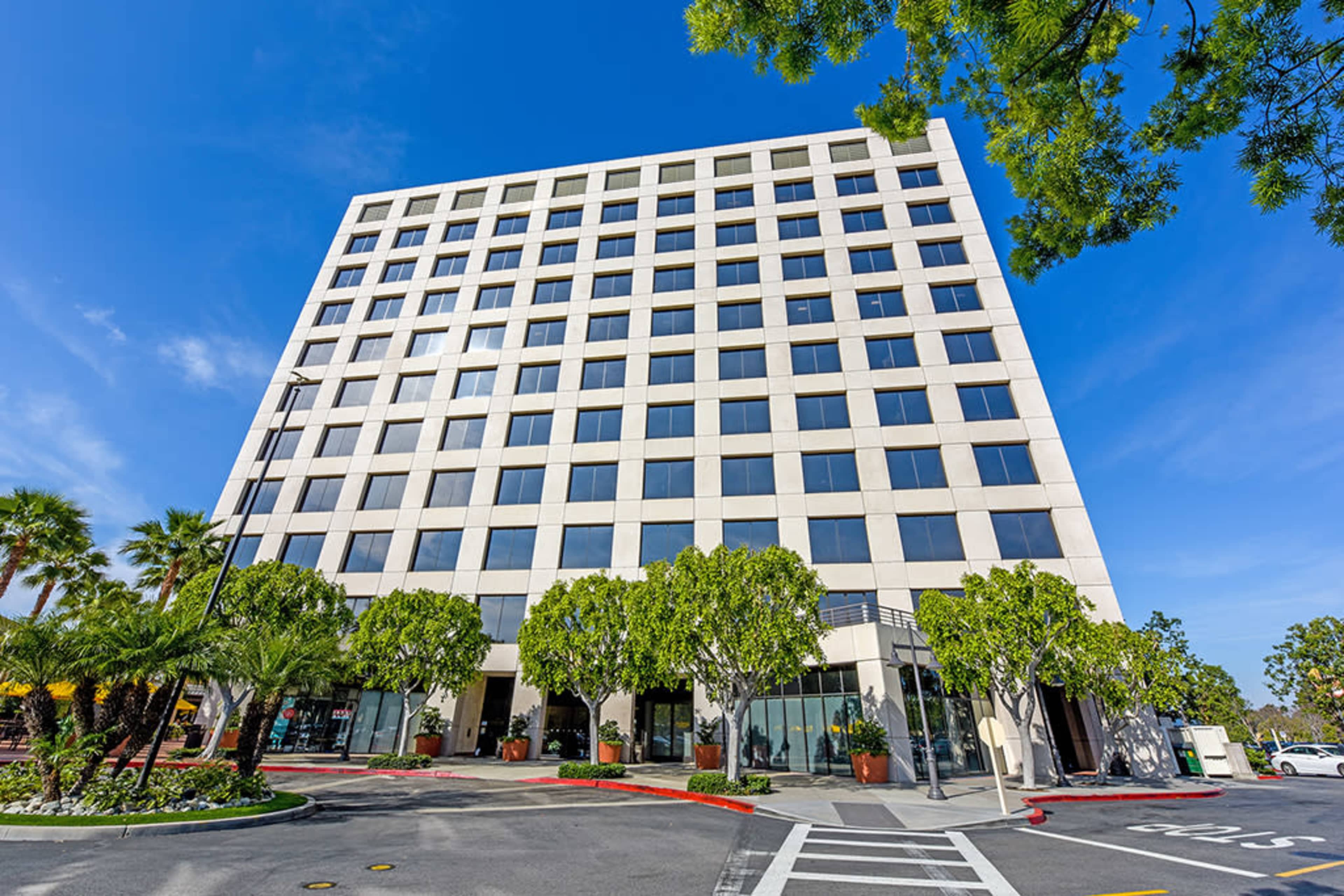 A modern, six-story office building with a flat roof and large windows is surrounded by landscaped greenery and palm trees.