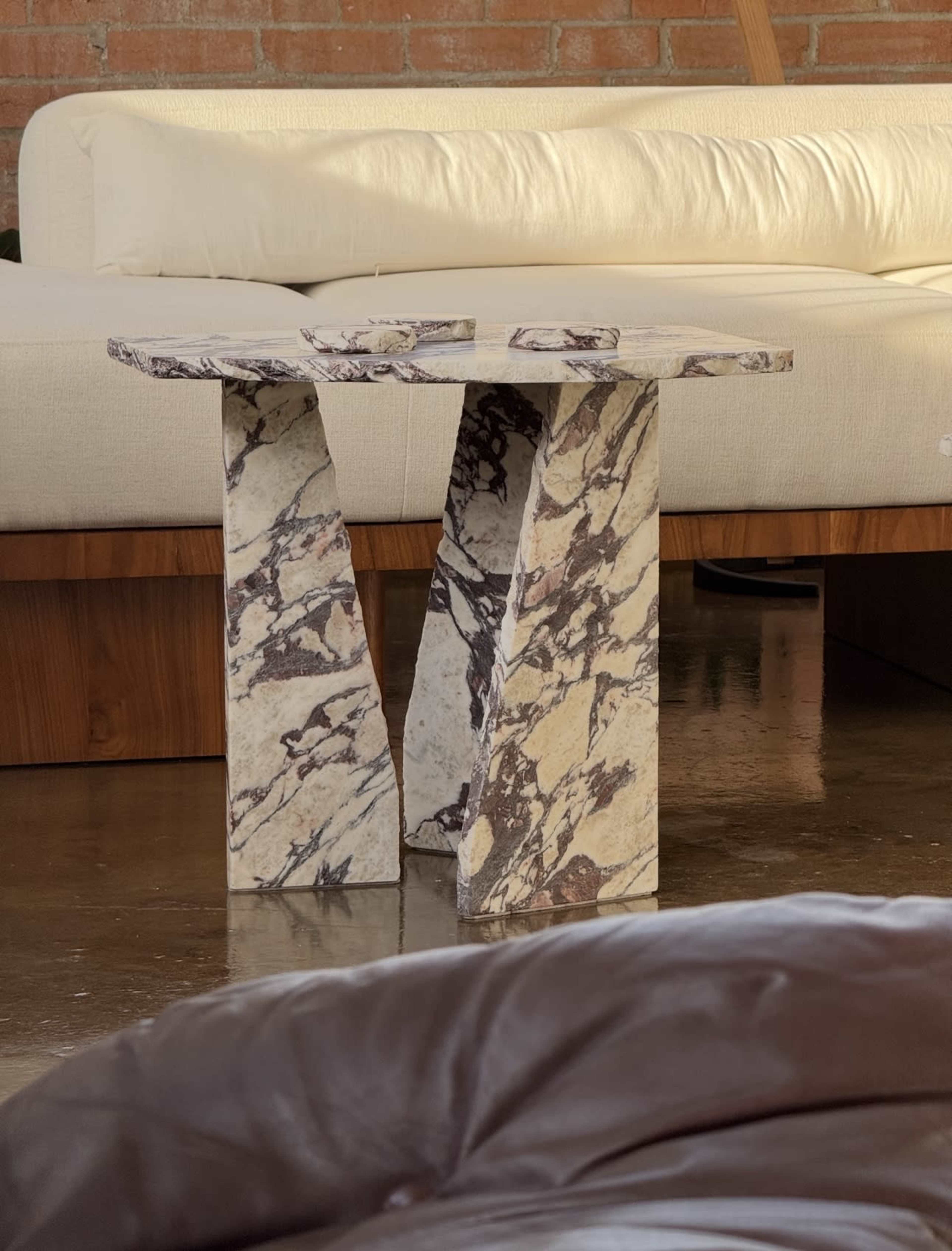 The image shows a marble side table with a unique, angular design placed in front of a light-colored sofa in a modern interior setting.