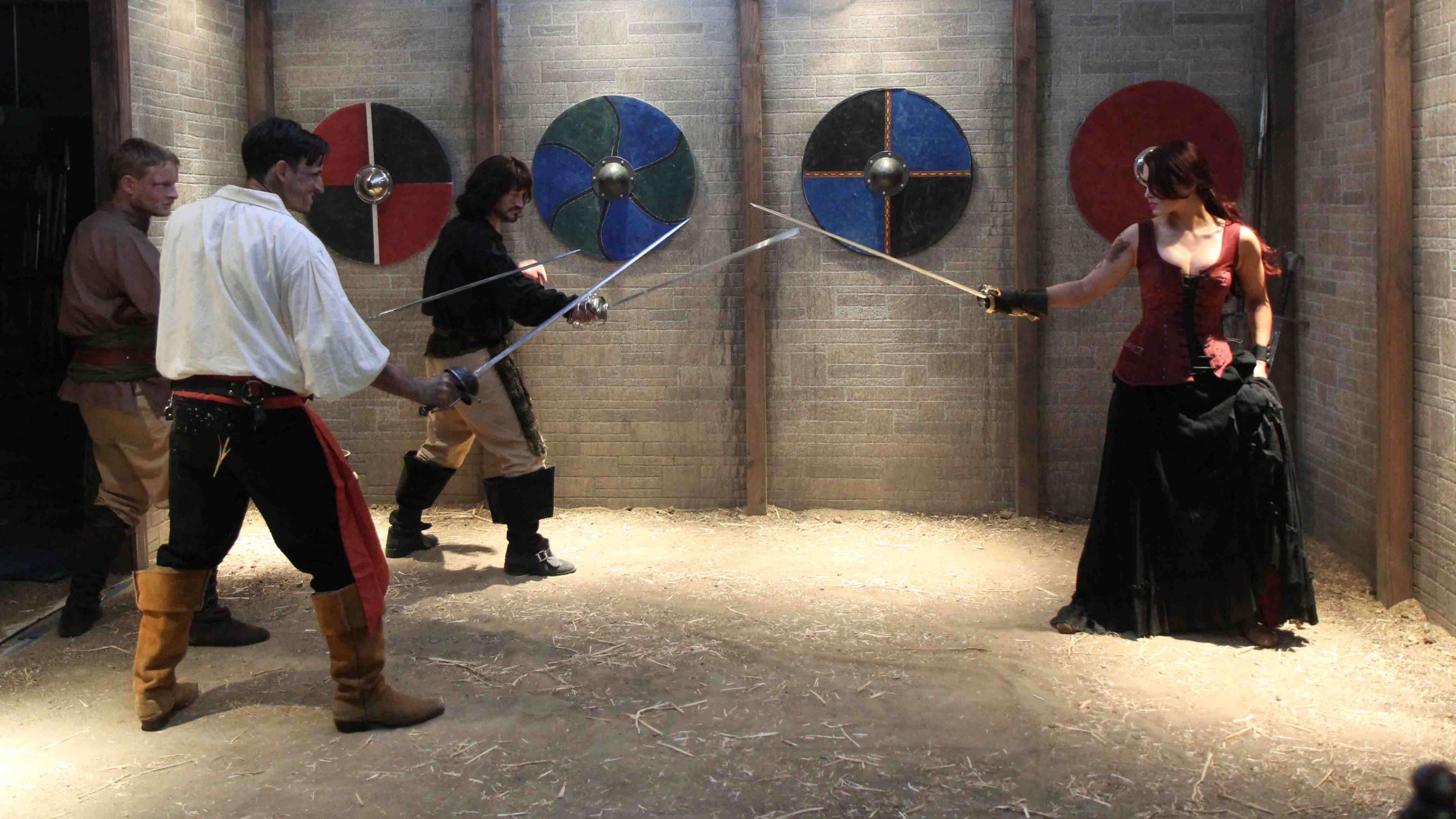 Four individuals are engaged in a sword fight in a barn-like setting, with brightly colored shields mounted on the walls behind them.