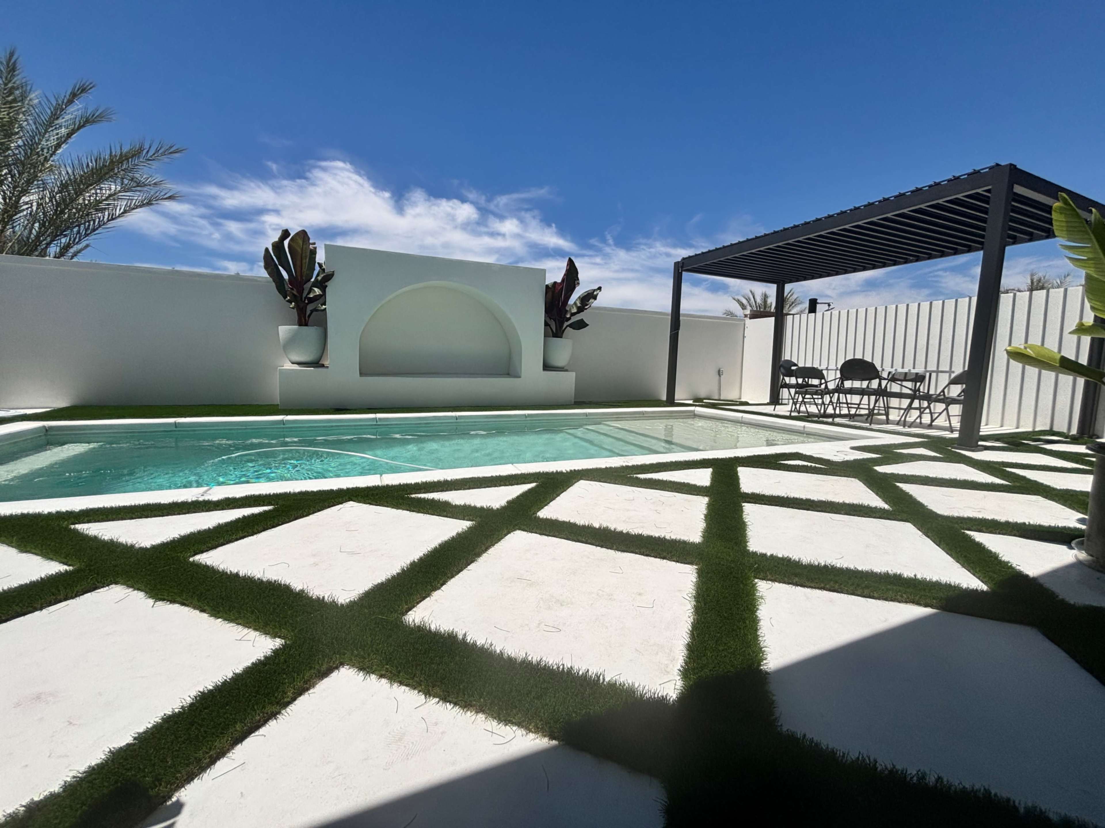 The image features a swimming pool surrounded by a geometric pattern of white concrete and artificial grass, with a shaded seating area in the background.