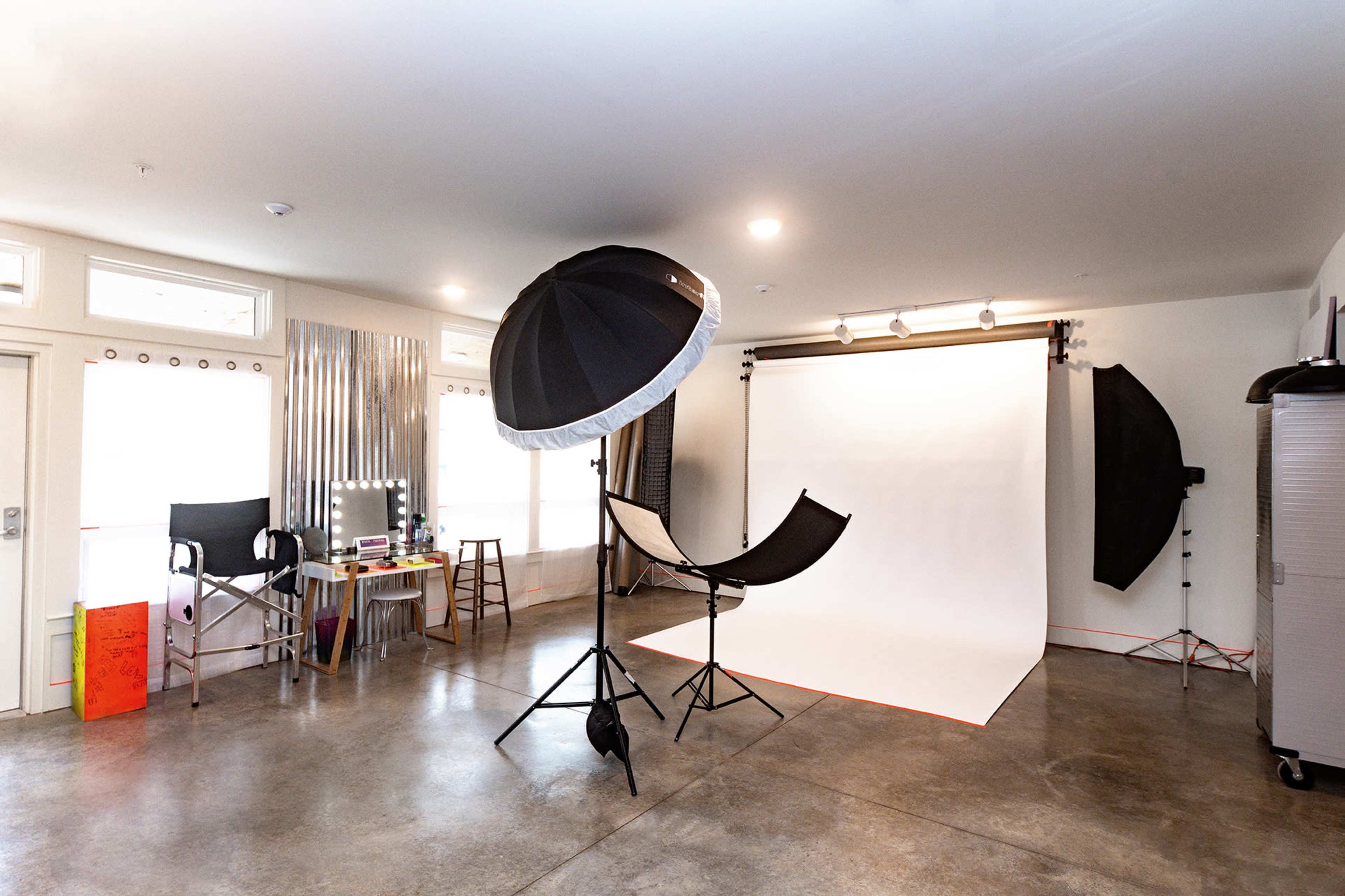 The image shows a photography studio featuring a white backdrop, a large umbrella light, and several pieces of equipment arranged around the space.