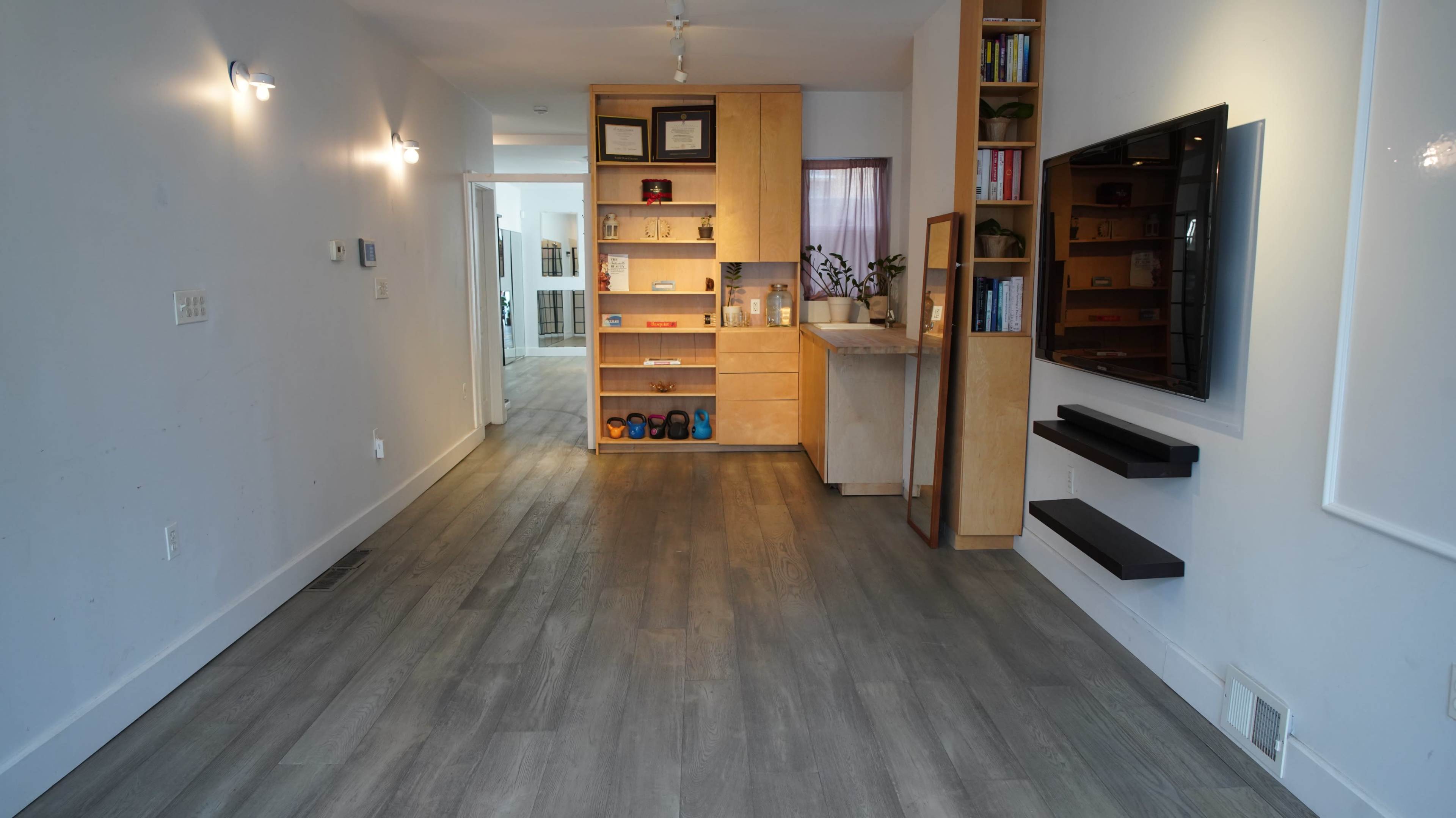 The image shows a modern, open interior space featuring a wooden shelving unit and a kitchen area, with gray flooring and minimalistic decor.