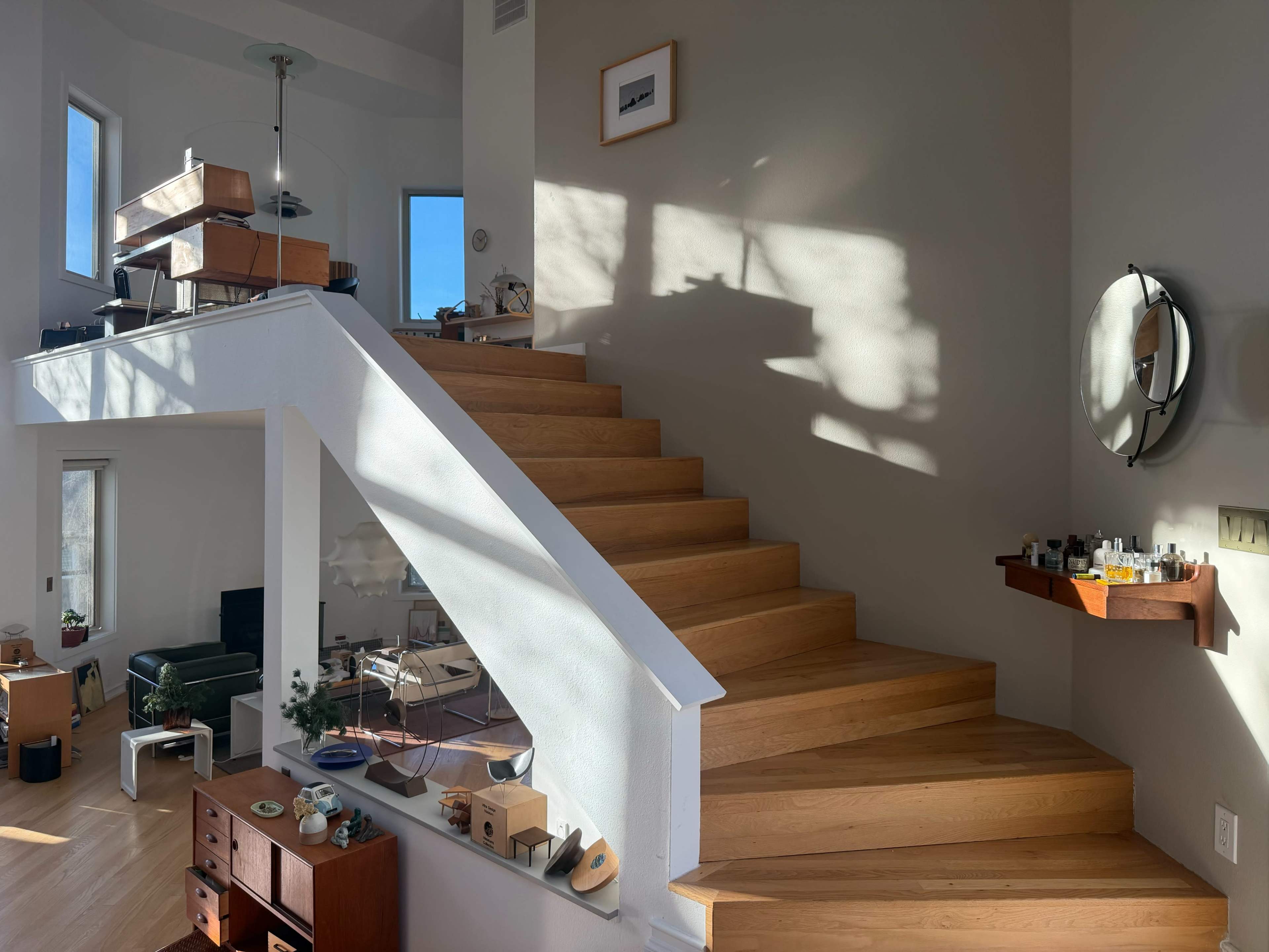 The image shows a wooden staircase leading to an upper level in a brightly lit modern living space, with furniture and decor visible in the background.