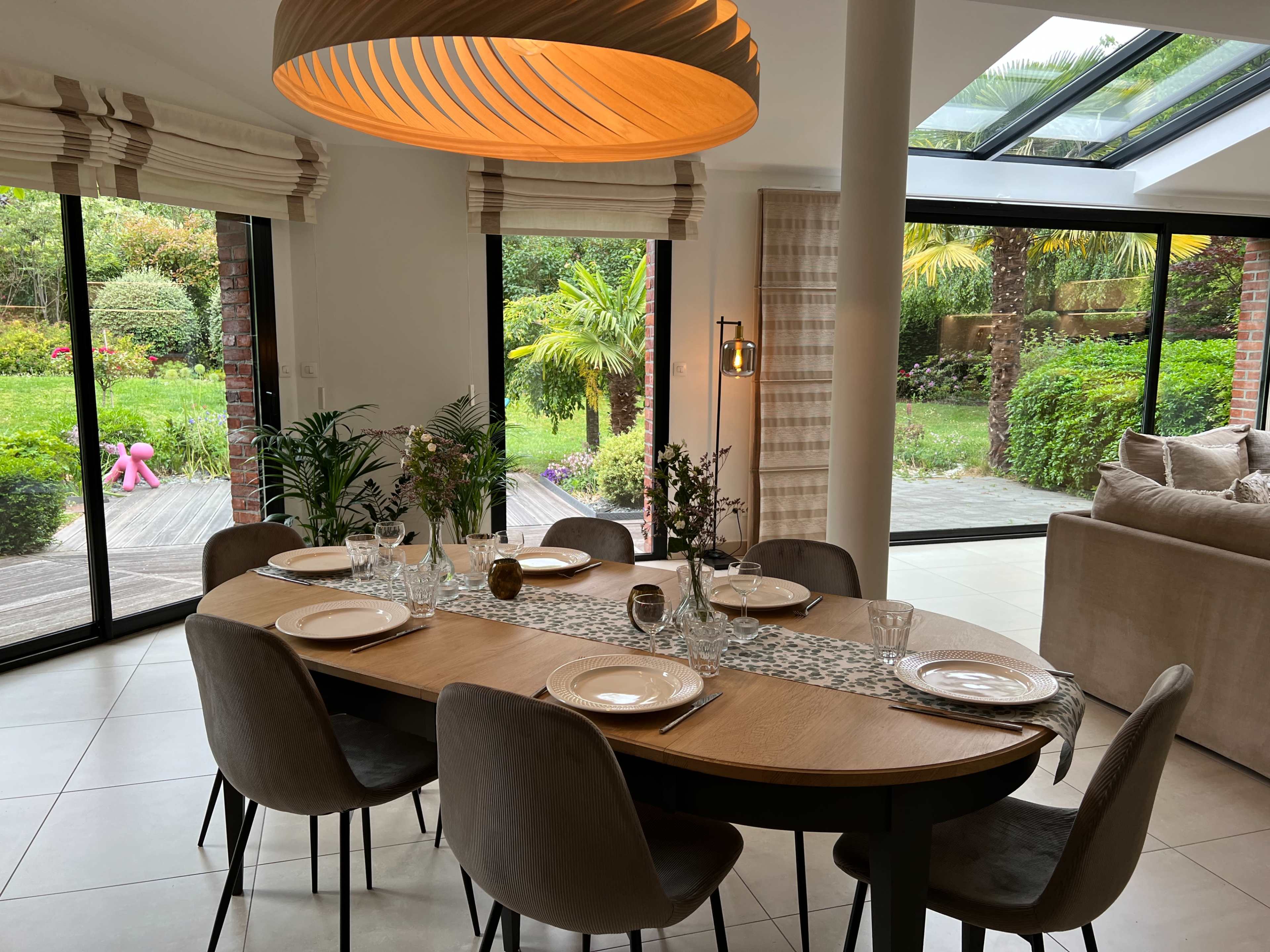 A modern dining area features a circular wooden table set with plates and glassware, surrounded by upholstered chairs, with large windows offering a view of a lush garden.