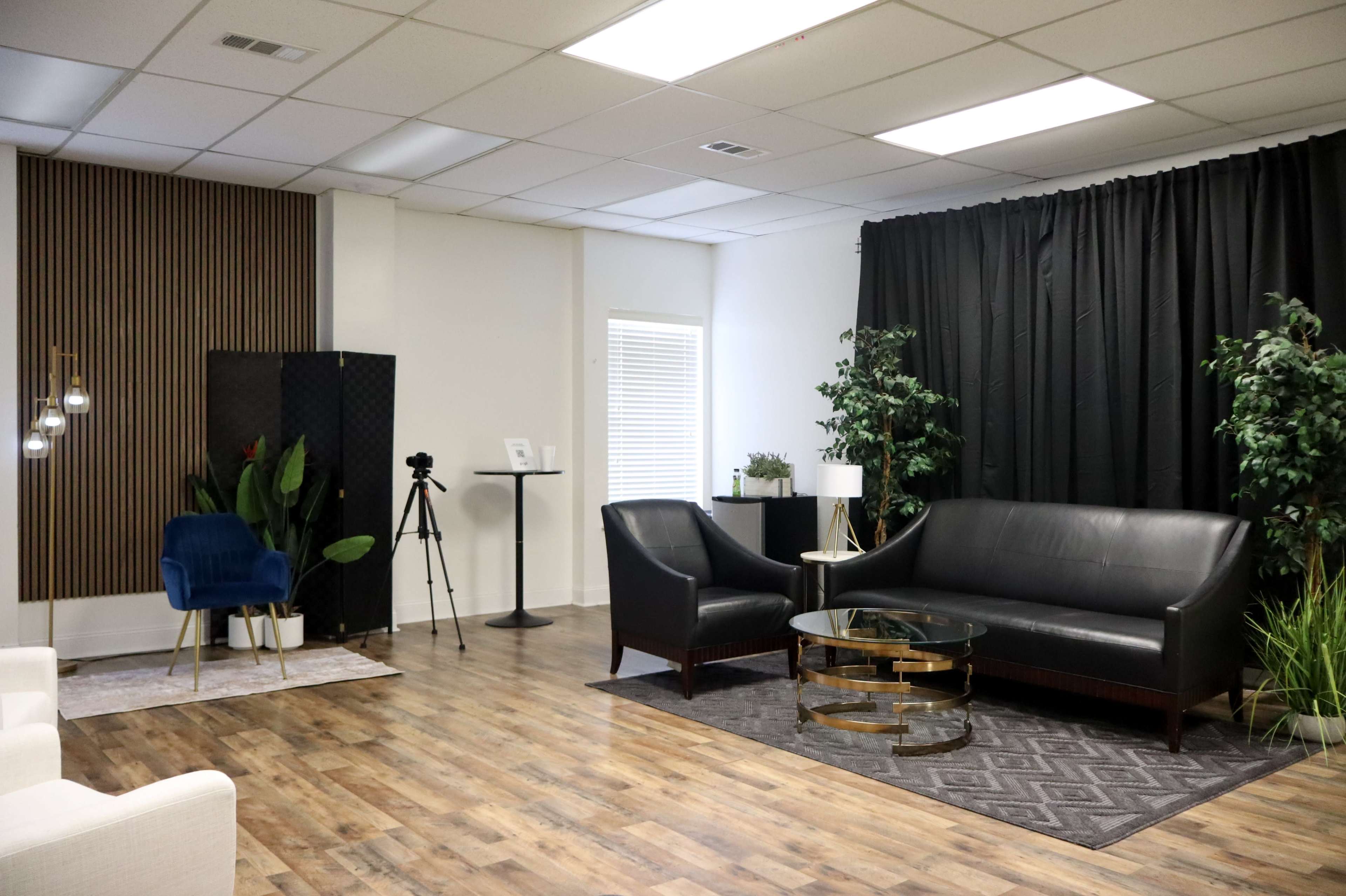 The image shows a modern indoor space with two black leather sofas, a glass coffee table, plants, and a backdrop of dark curtains, alongside a tripod and lighting setup.