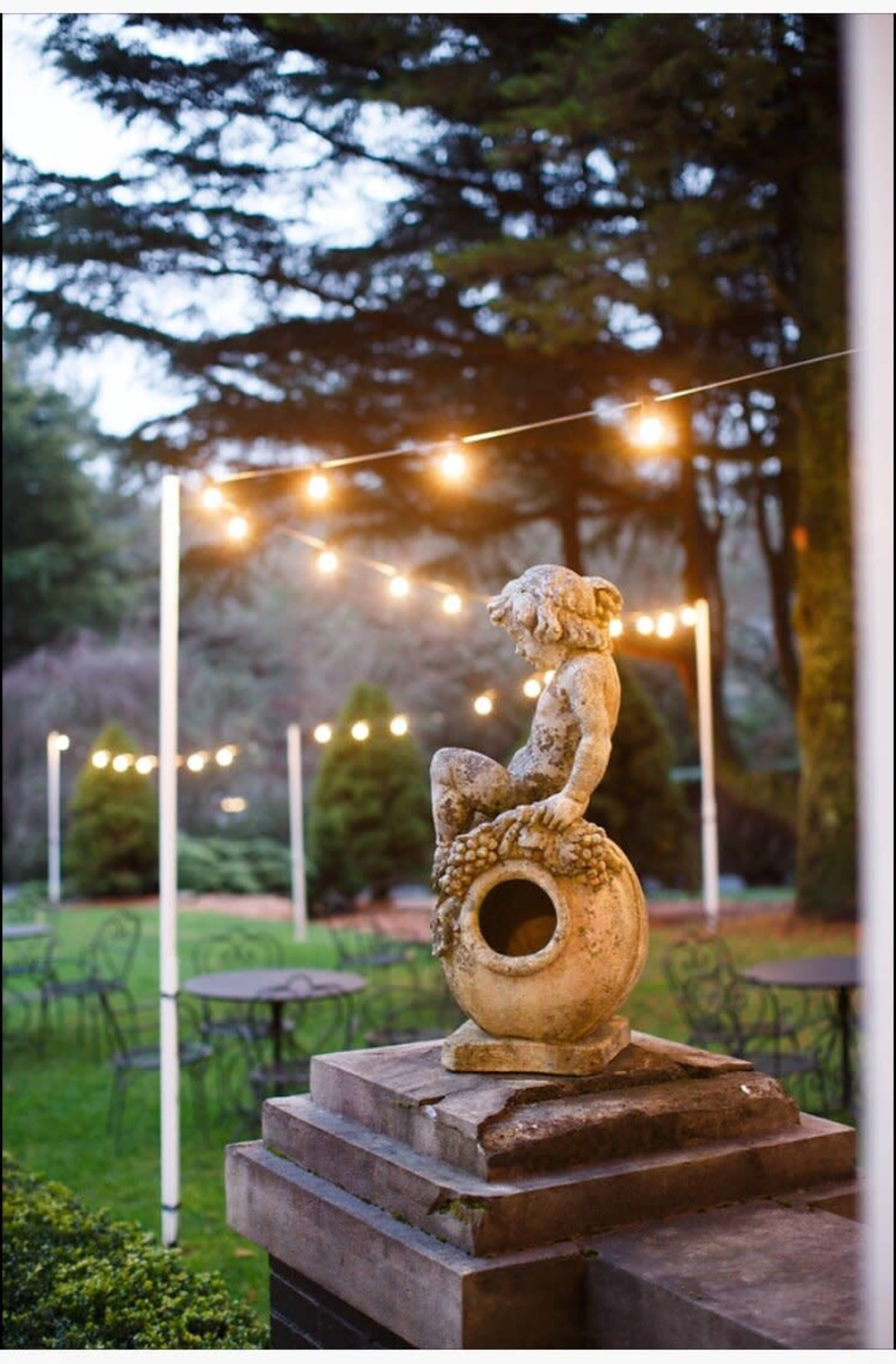 A stone statue of a cherub sits atop a pedestal in a garden illuminated by string lights.