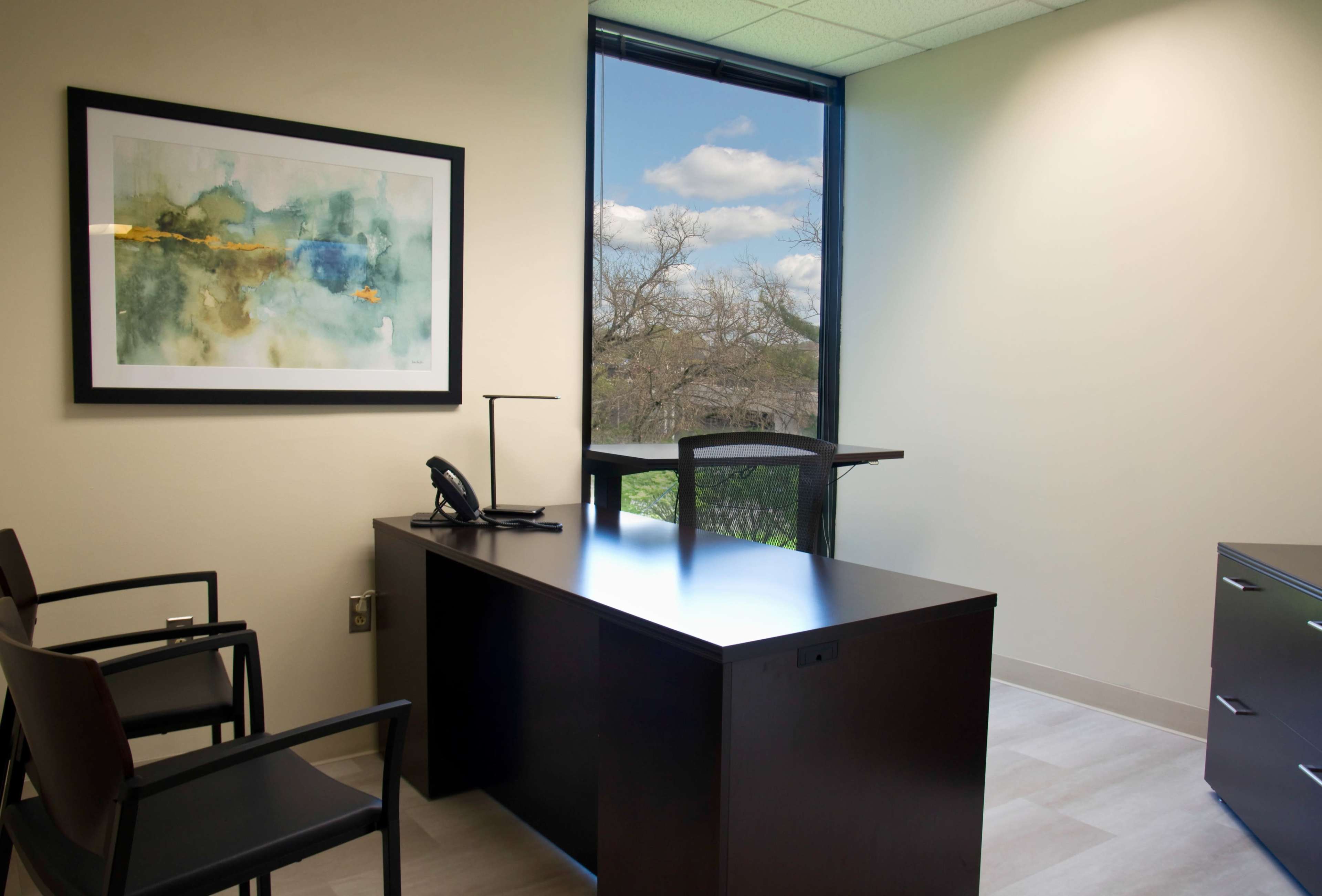The image shows a modern office space with a desk, a phone, two chairs, and a window offering a view of trees and a blue sky.