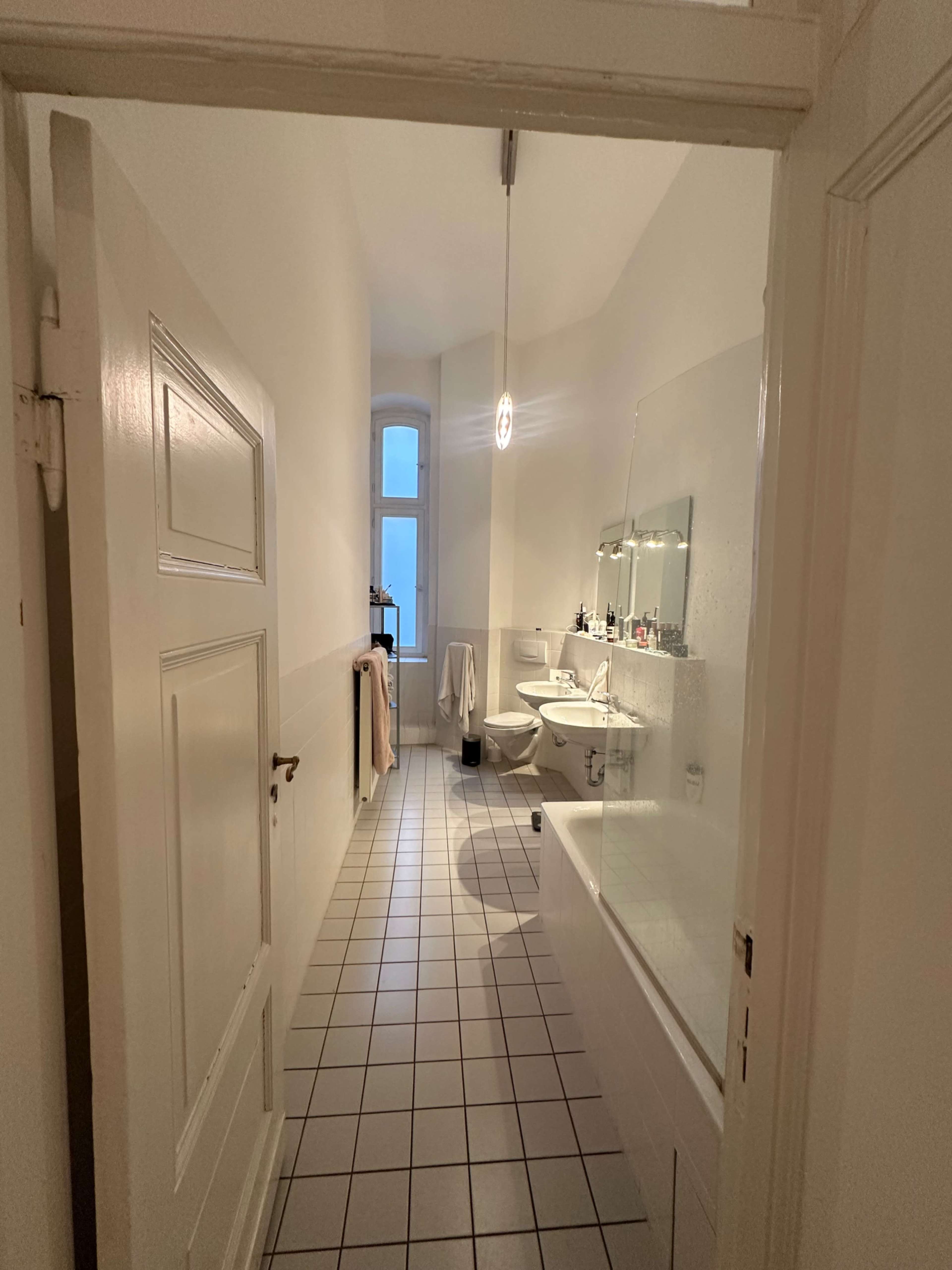 The image shows a bathroom viewed from an open doorway, featuring a bathtub, sink, and tiled floor, with a window allowing natural light.