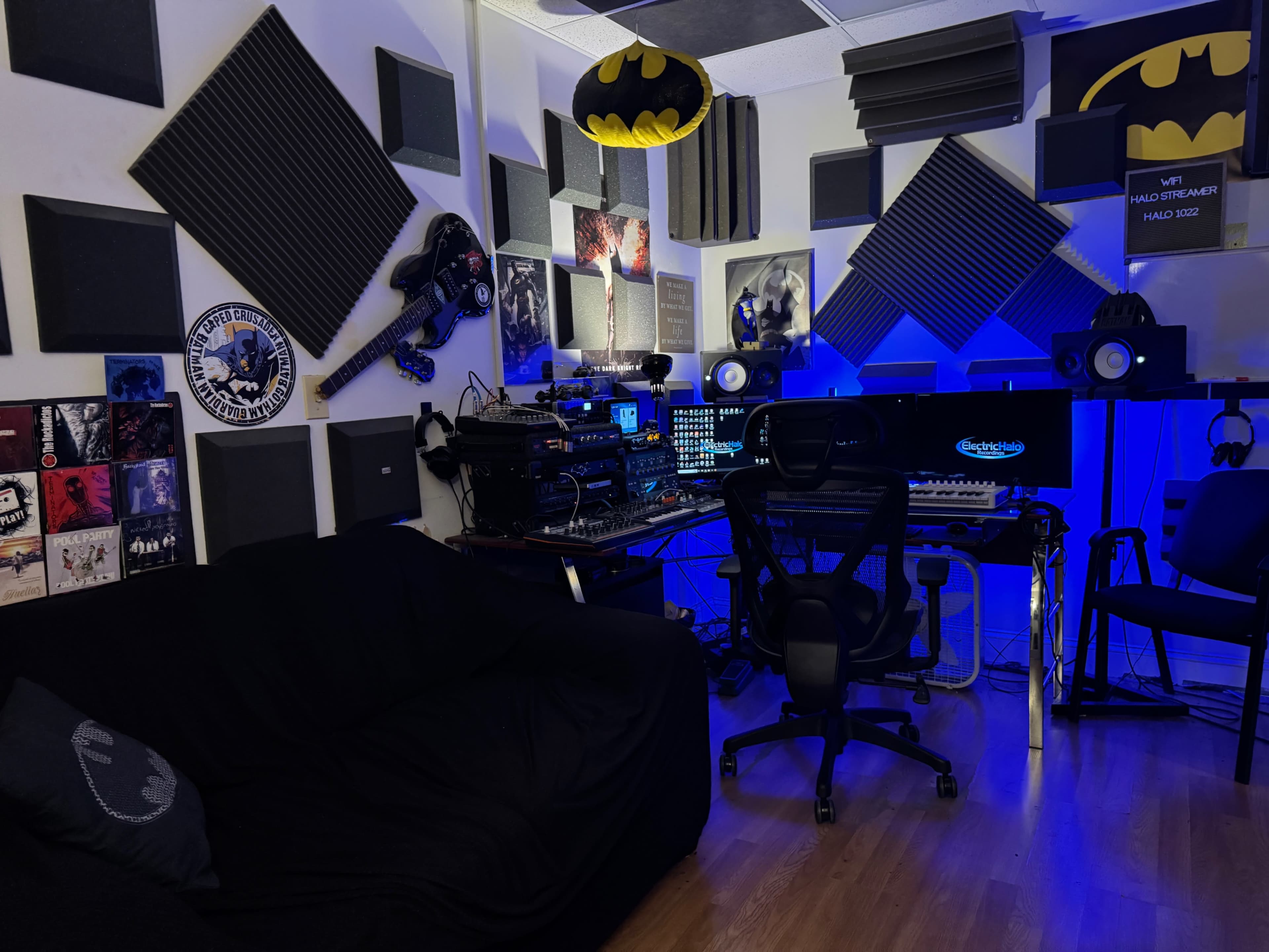 The image shows a music production studio with soundproofing panels on the walls, a keyboard, speakers, and various musical equipment, along with a black couch and a Batman-themed decoration.